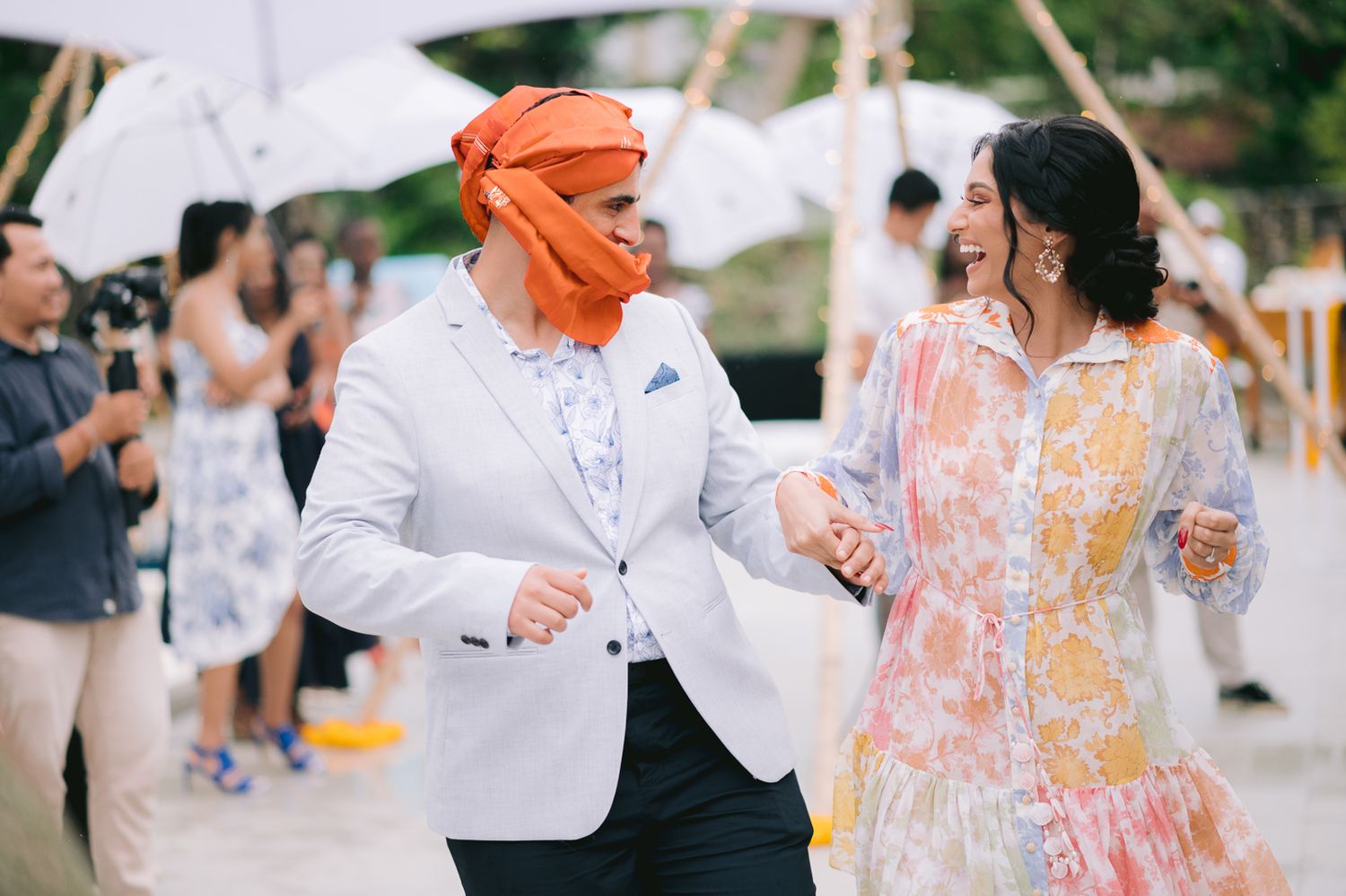 Two people dancing joyfully outdoors, one wearing an orange turban and white blazer, the other in a floral dress.