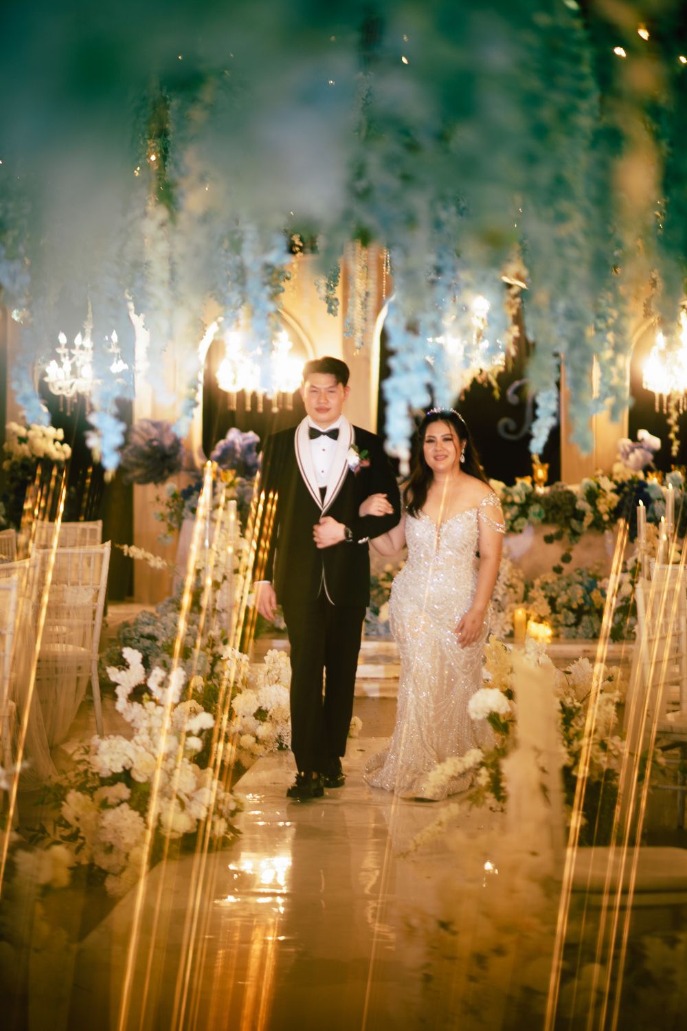 Bride and groom walk down a sparkling aisle surrounded by lush floral arrangements and golden light at elegant wedding reception.