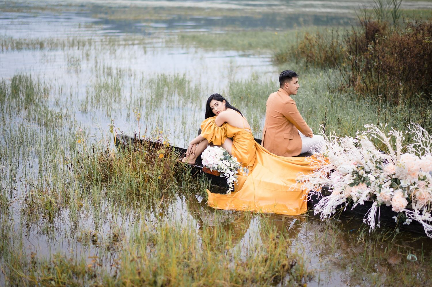 Couple in orange outfits sitting in a rustic boat surrounded by wildflowers in a serene marshy landscape.