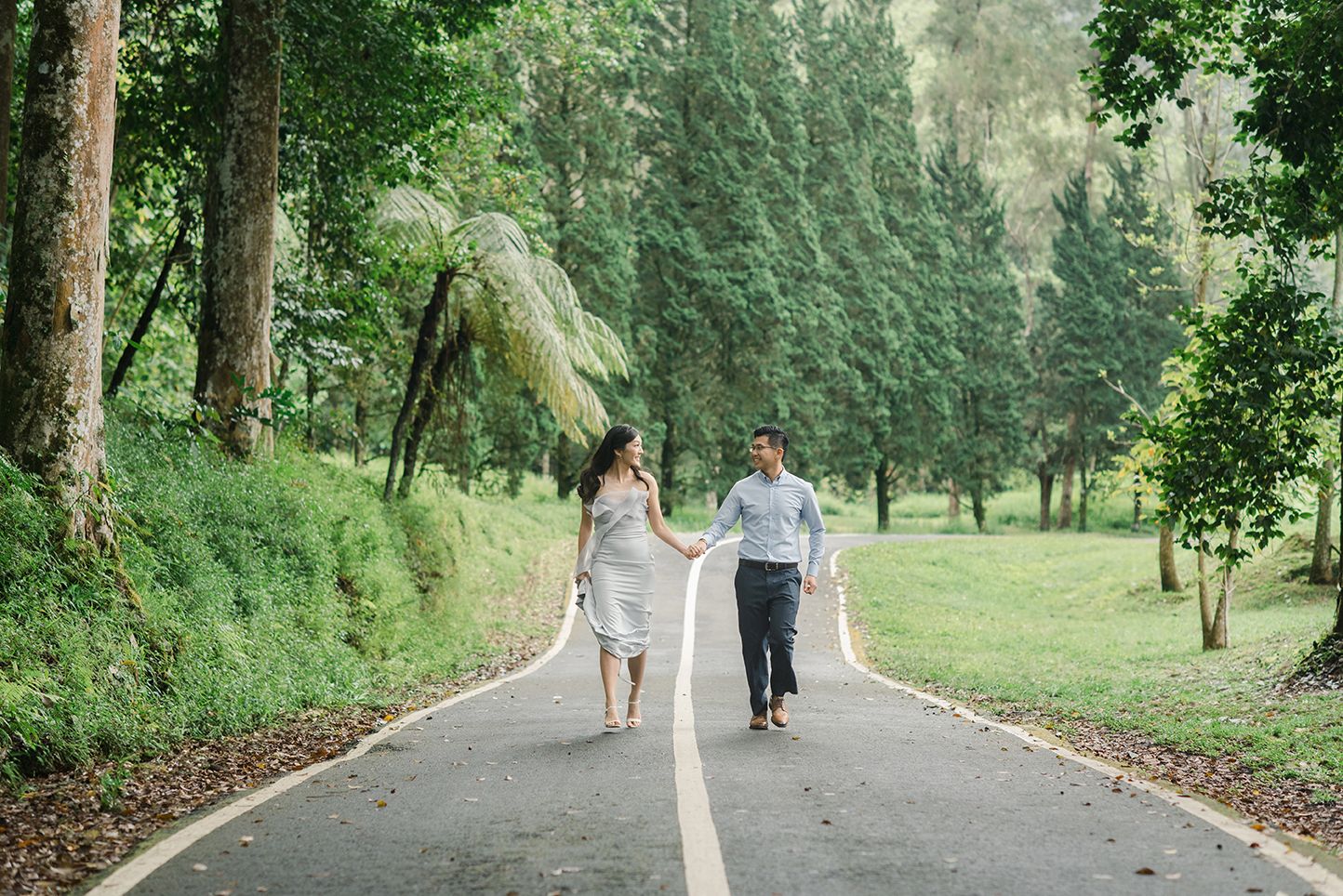 Couple holding hands walking down a tree-lined road surrounded by lush green forest.
