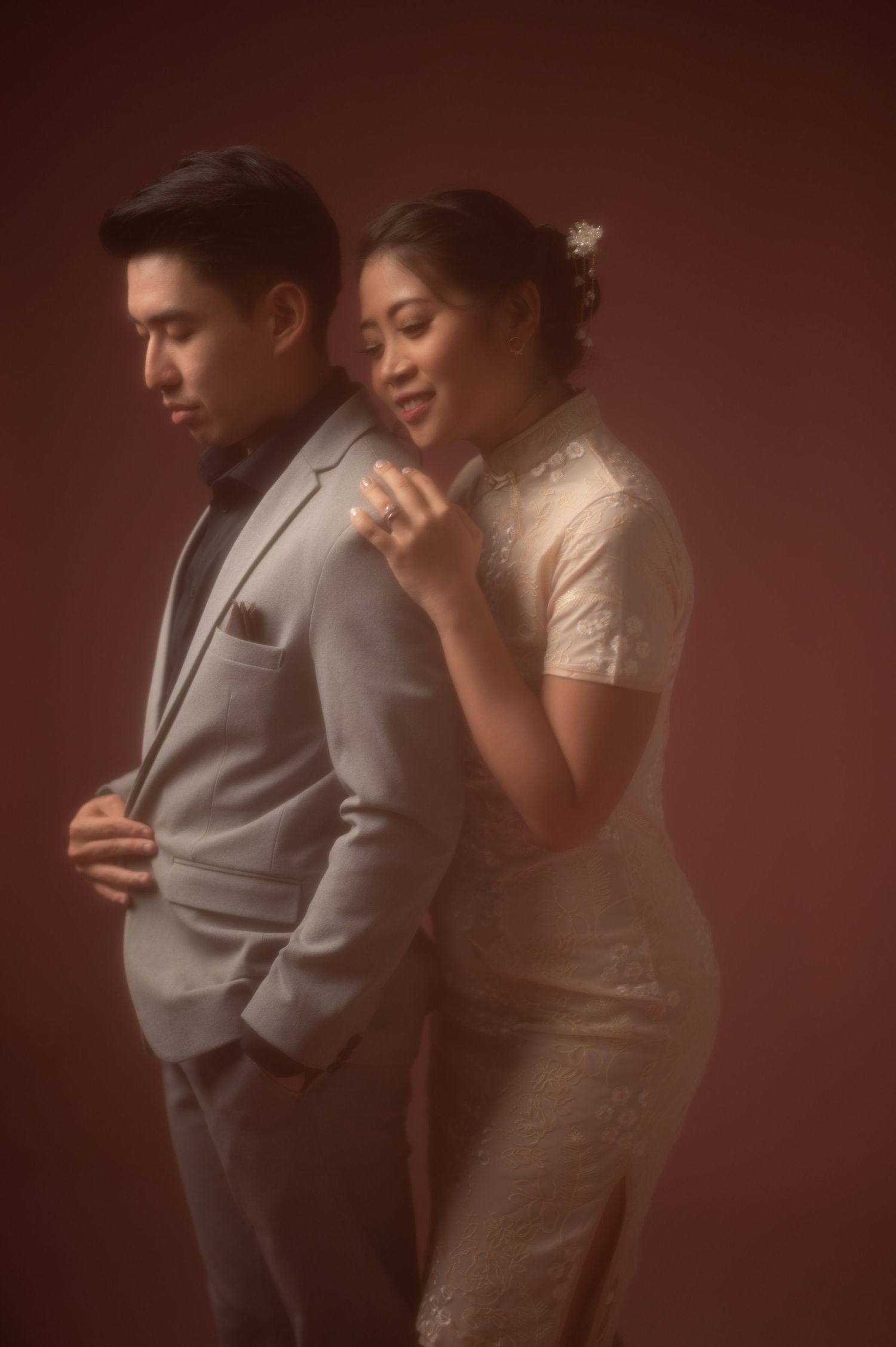 Elegant couple in formal attire pose together, woman in lace qipao embracing man in gray suit against moody red backdrop.