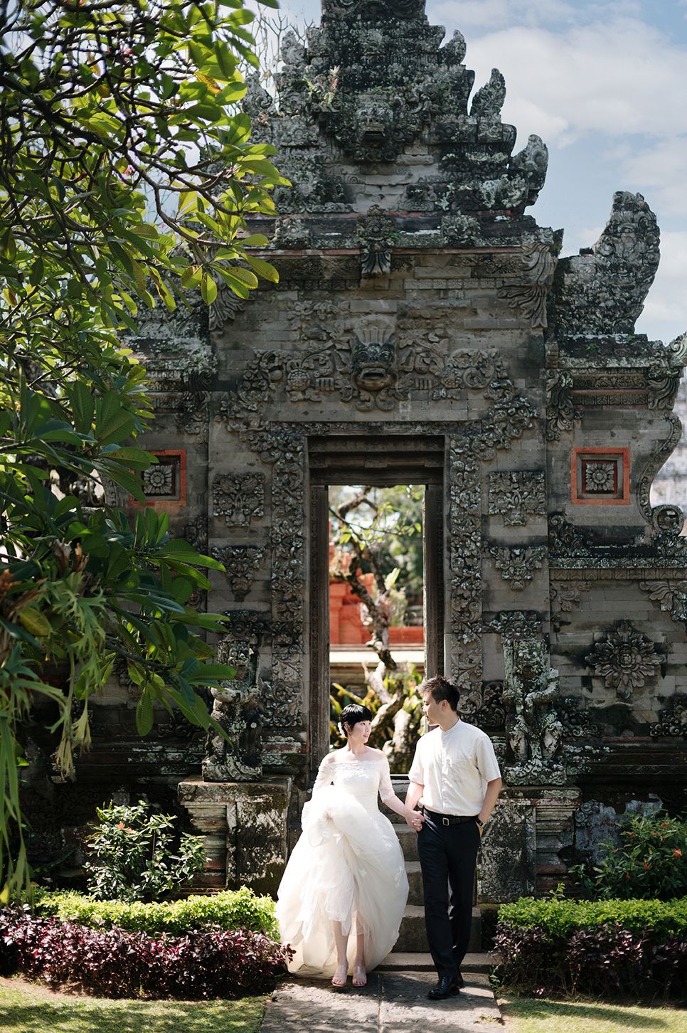 Couple in wedding attire poses before ornate Balinese stone temple gate surrounded by lush tropical gardens.
