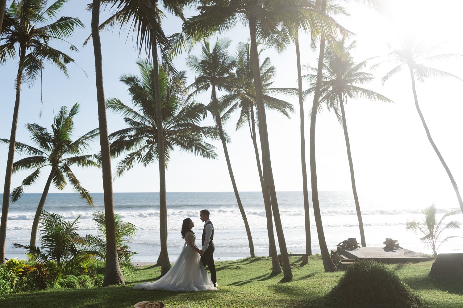 Bride and groom stand among tall palm trees overlooking a tropical beach at sunset during their destination wedding.