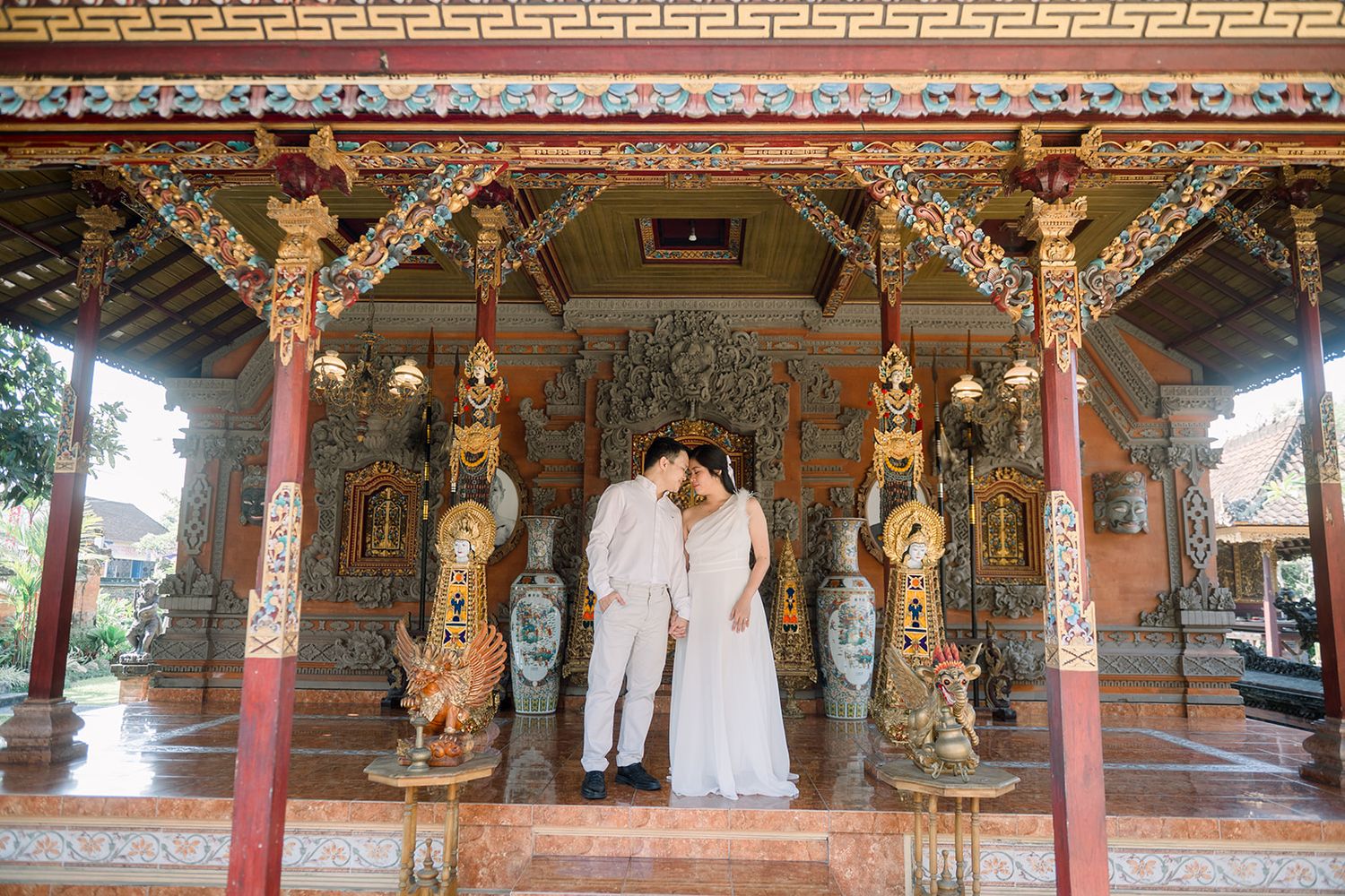 Couple in white attire sharing a romantic moment at an ornate Balinese temple with red columns and gold decor.