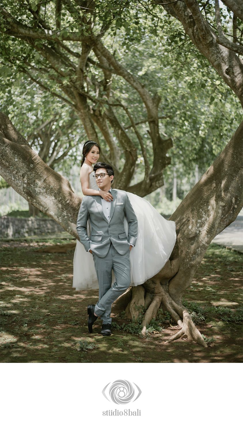 Couple poses romantically under sprawling trees, groom in gray suit, bride in white dress, for a dreamy outdoor pre-wedding shoot.