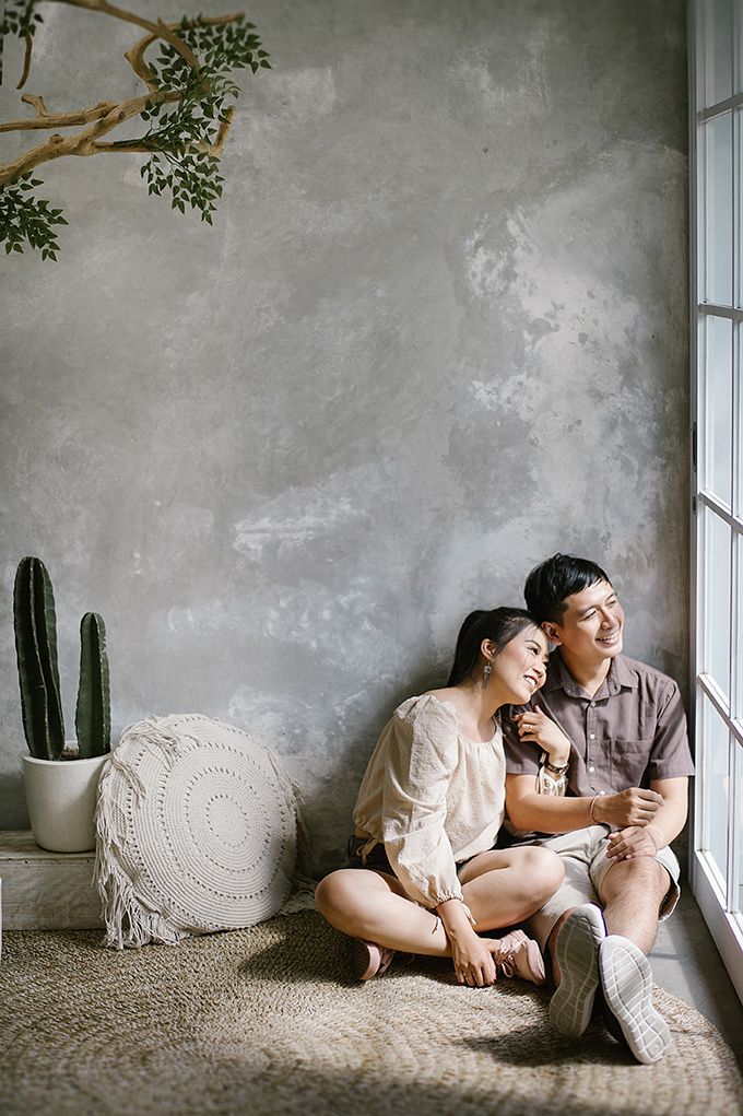 A couple sits together on the floor against a gray wall, smiling warmly near a window with a cactus nearby.