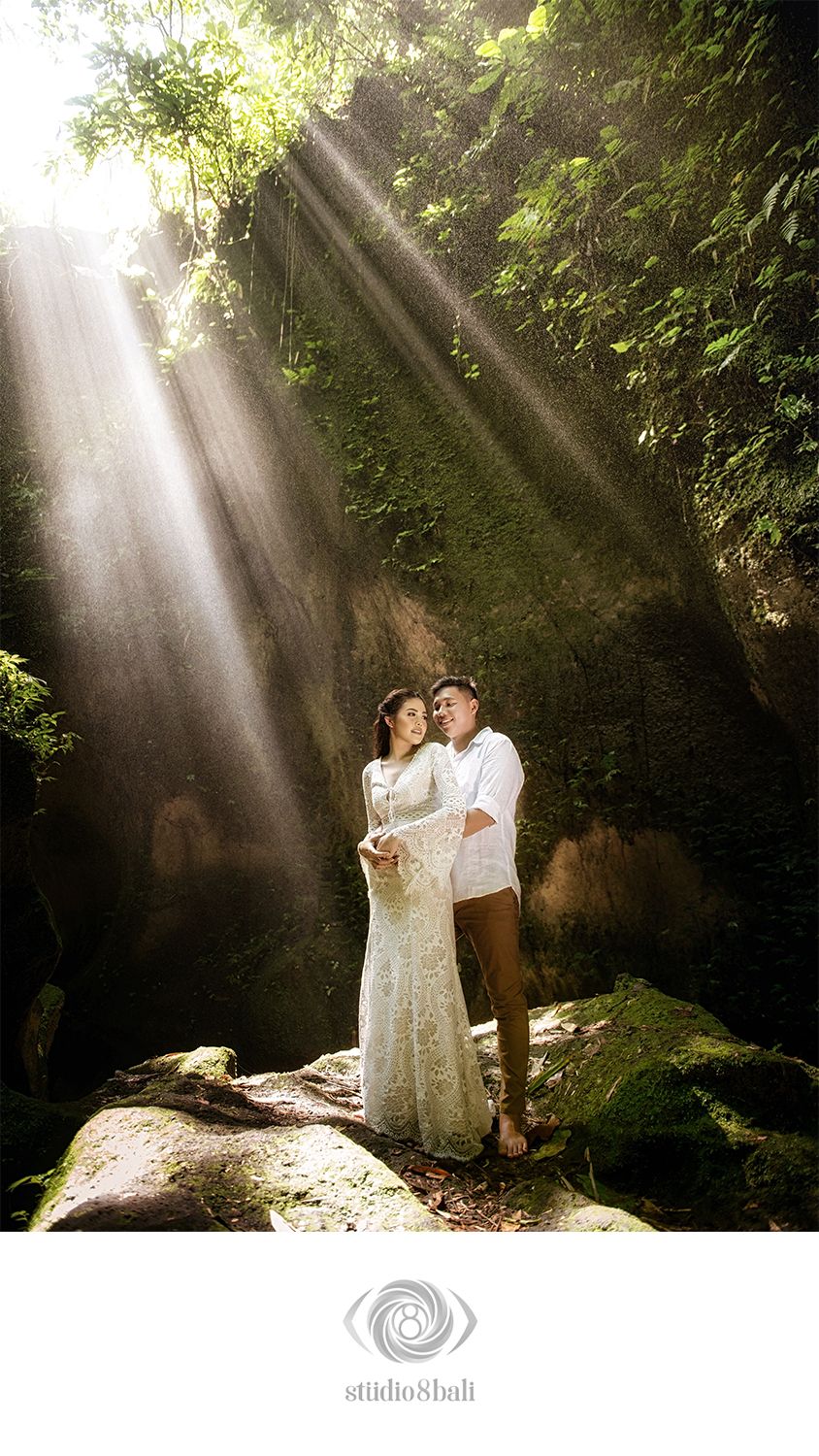 Couple embracing in lush forest canyon with dramatic sunbeams, woman in white lace gown, professional wedding photography by Studio 8 Bali.