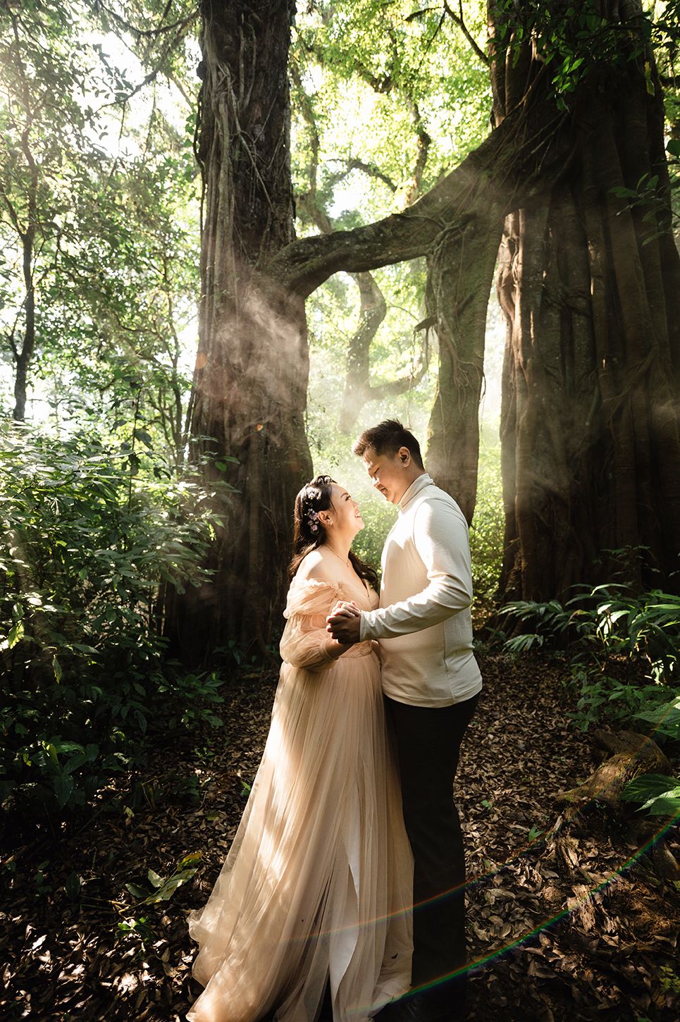 Couple embracing in enchanted forest with sunbeams filtering through ancient trees, bride in flowing gown.