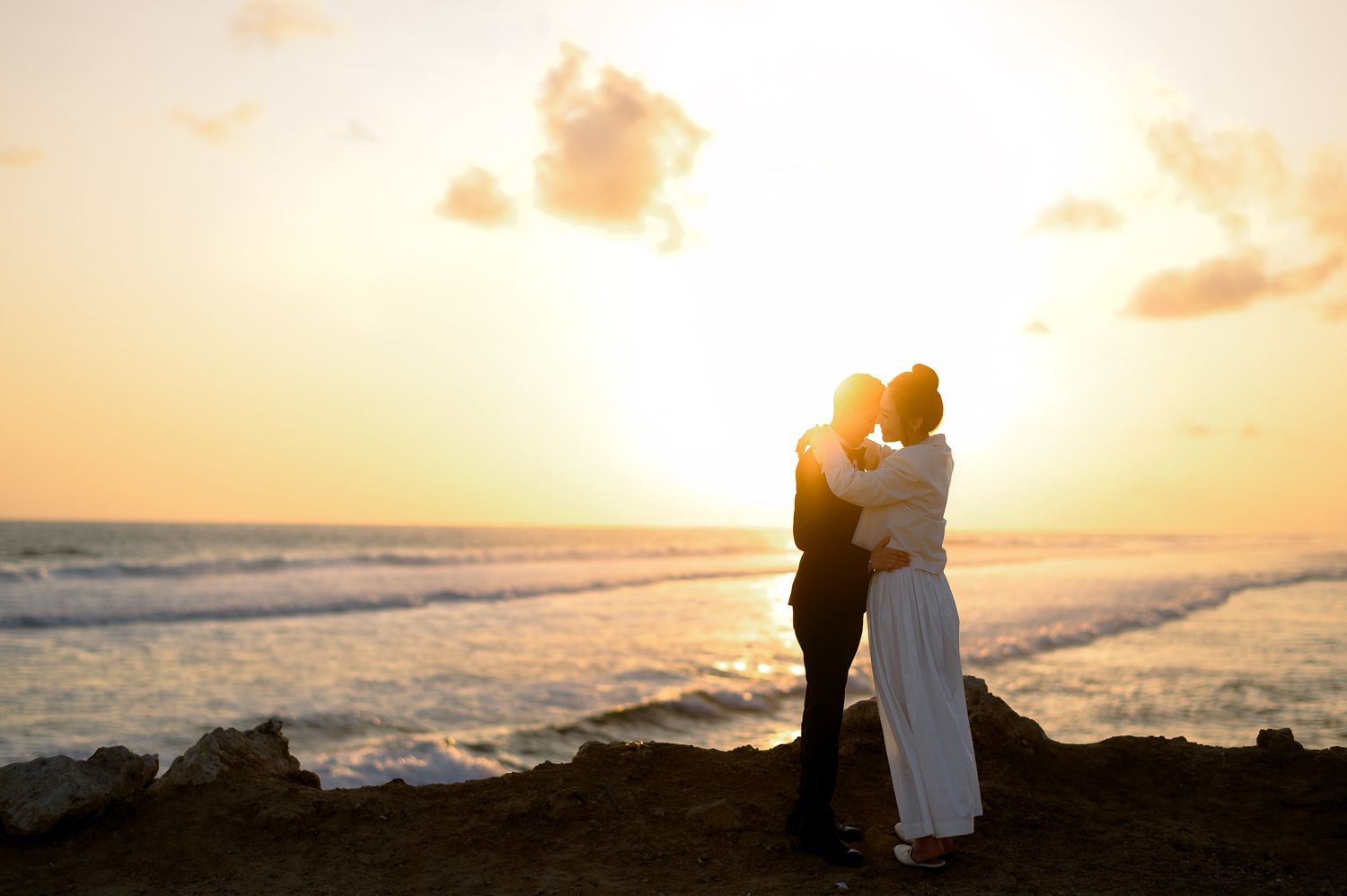 Couple embracing at sunset on rocky beach with golden ocean waves in background, romantic silhouette photo.