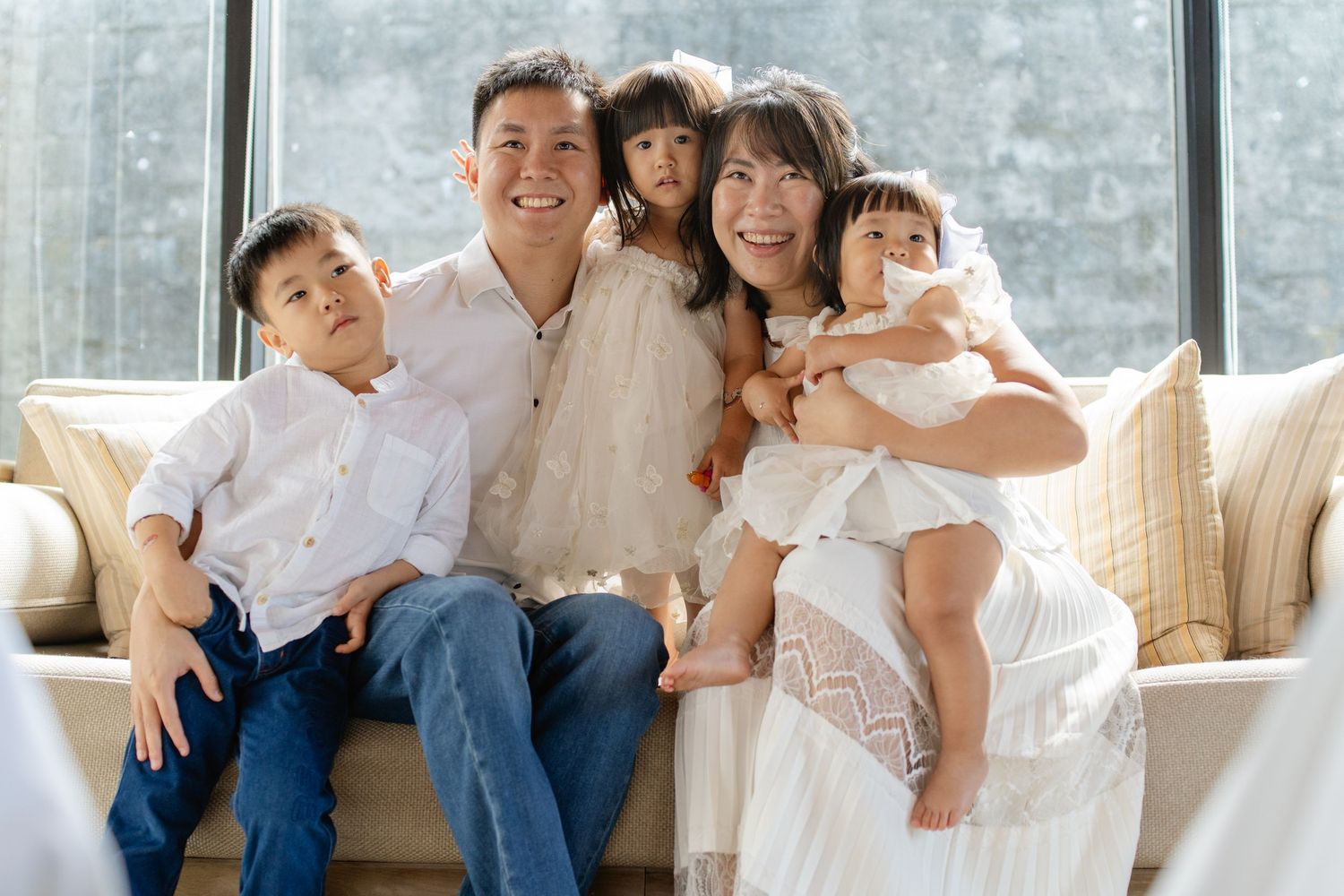 Happy Asian family of five posing together on a couch near large windows in coordinating white outfits.