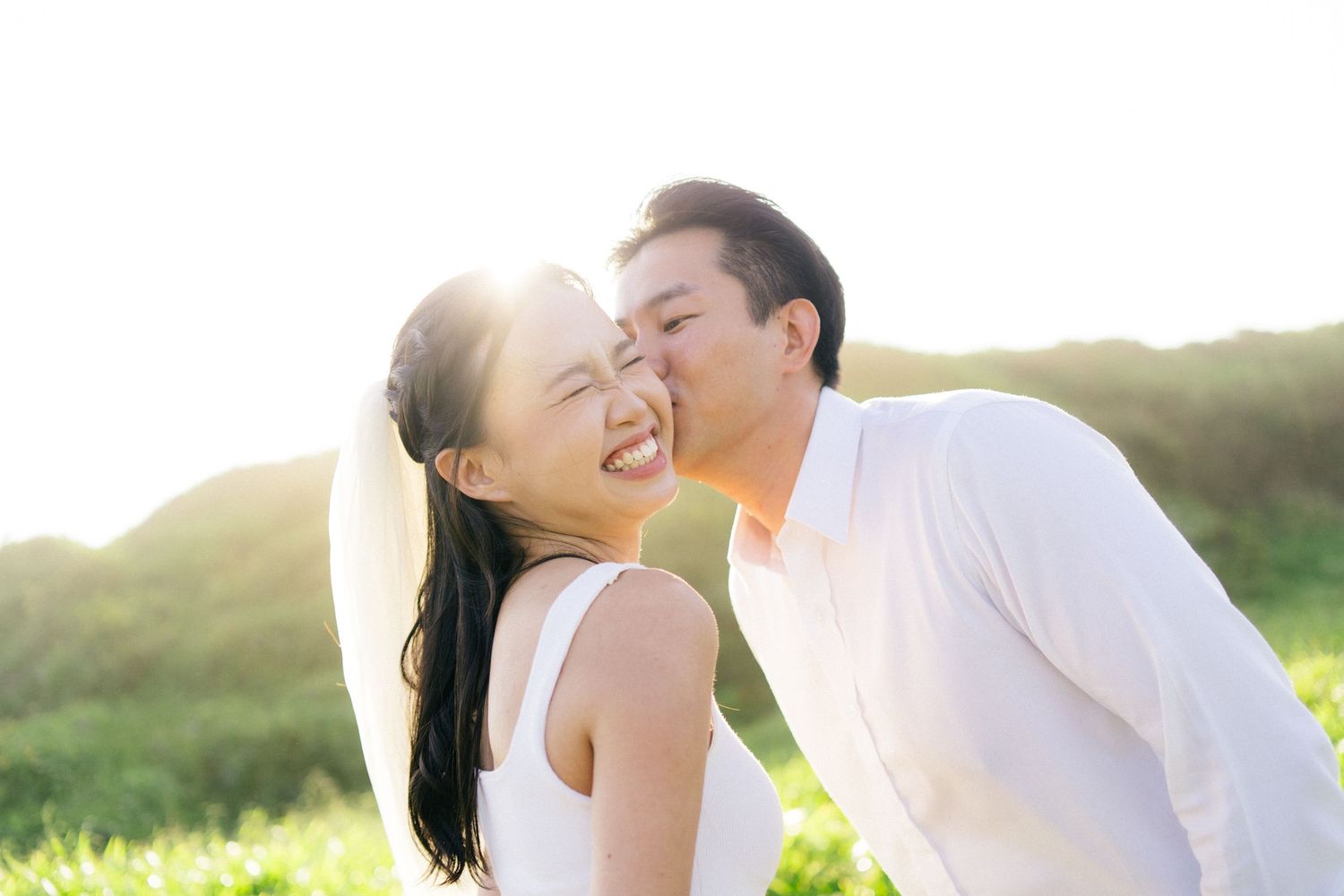 Couple sharing a romantic kiss outdoors in sunlit green field, man in white shirt kissing smiling woman.