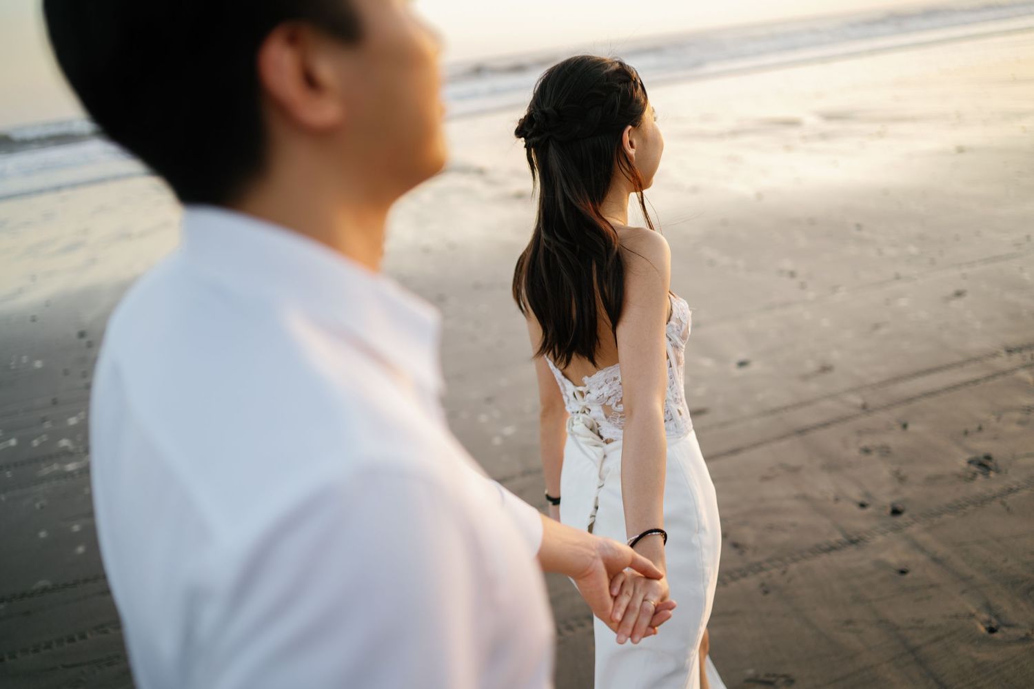 Bride in lace wedding gown holds groom's hand on sandy beach at sunset.