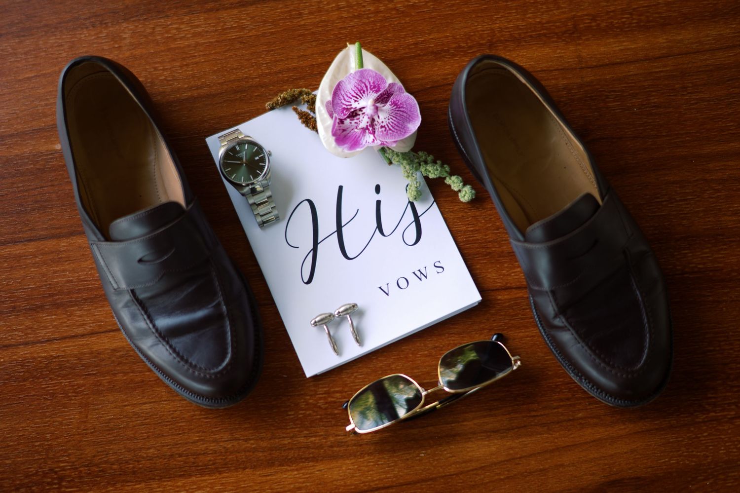 Groom's wedding flat lay with dark loafers, vow book, cufflinks, sunglasses, and floral accent on wood.