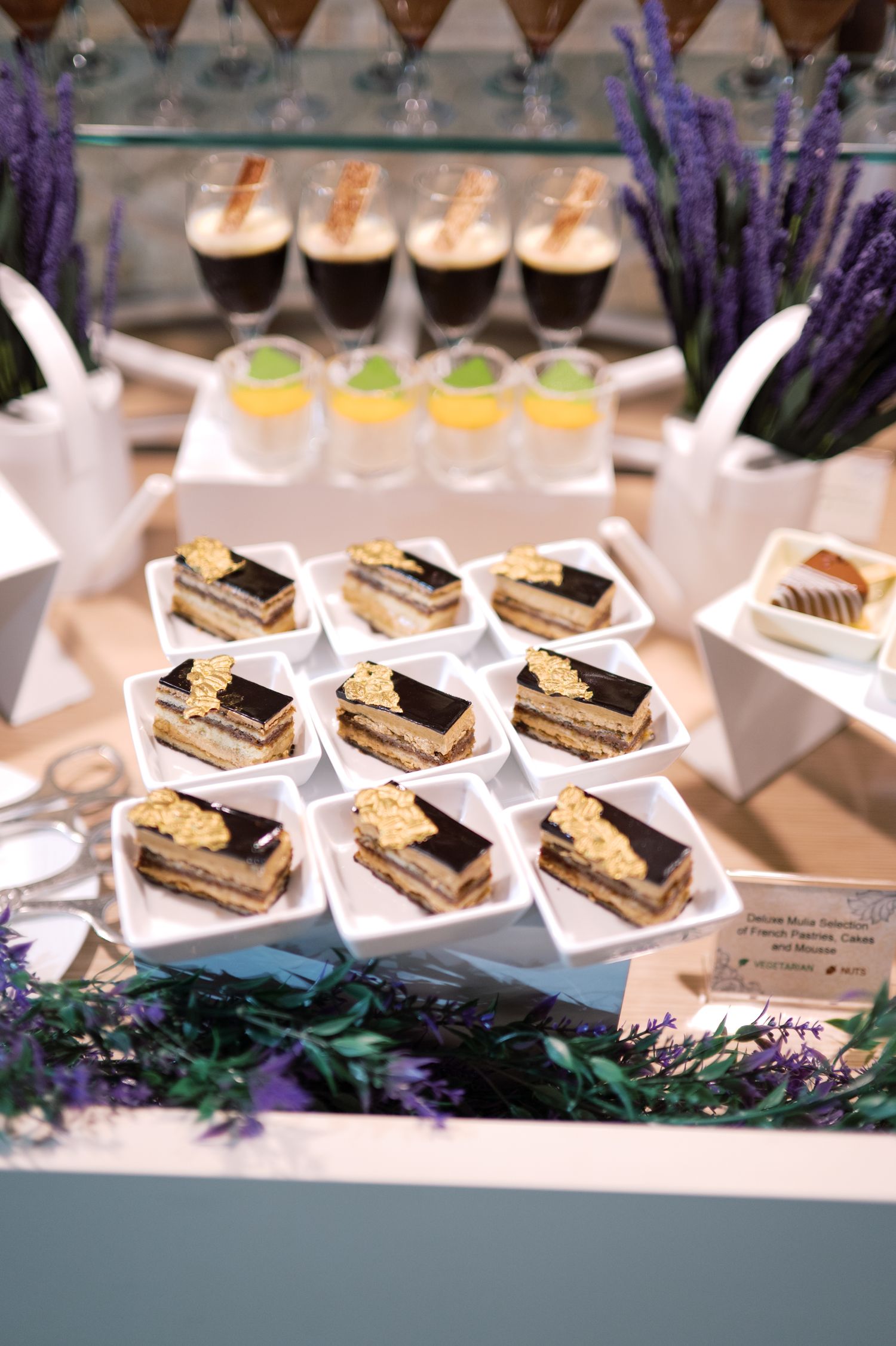 Elegant dessert display featuring chocolate opera cake slices on white plates with coffee drinks and lavender decor.