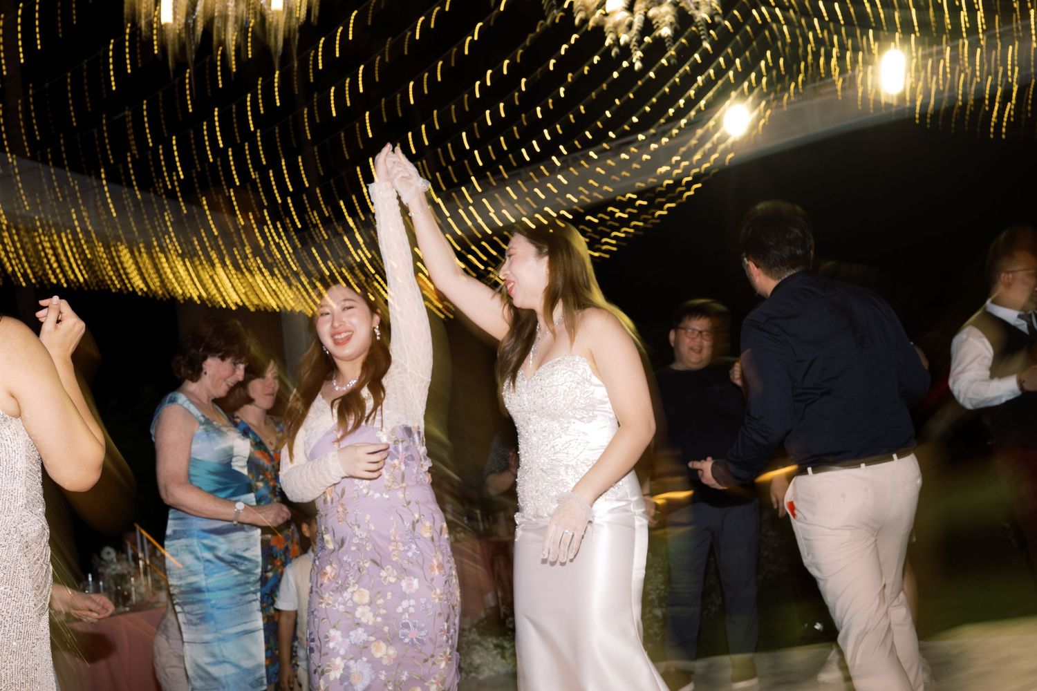 Two women dancing joyfully at a wedding reception under warm string lights.