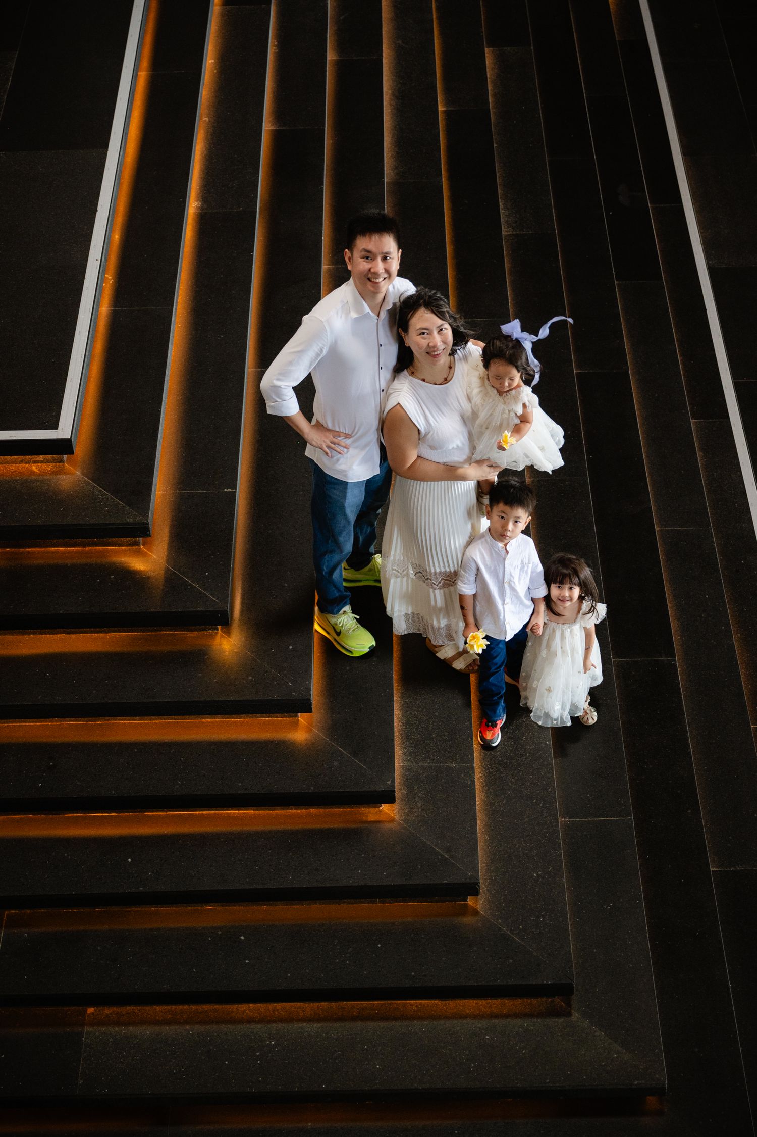 Family of five in white outfits posing on modern dark staircase, photographed from above.