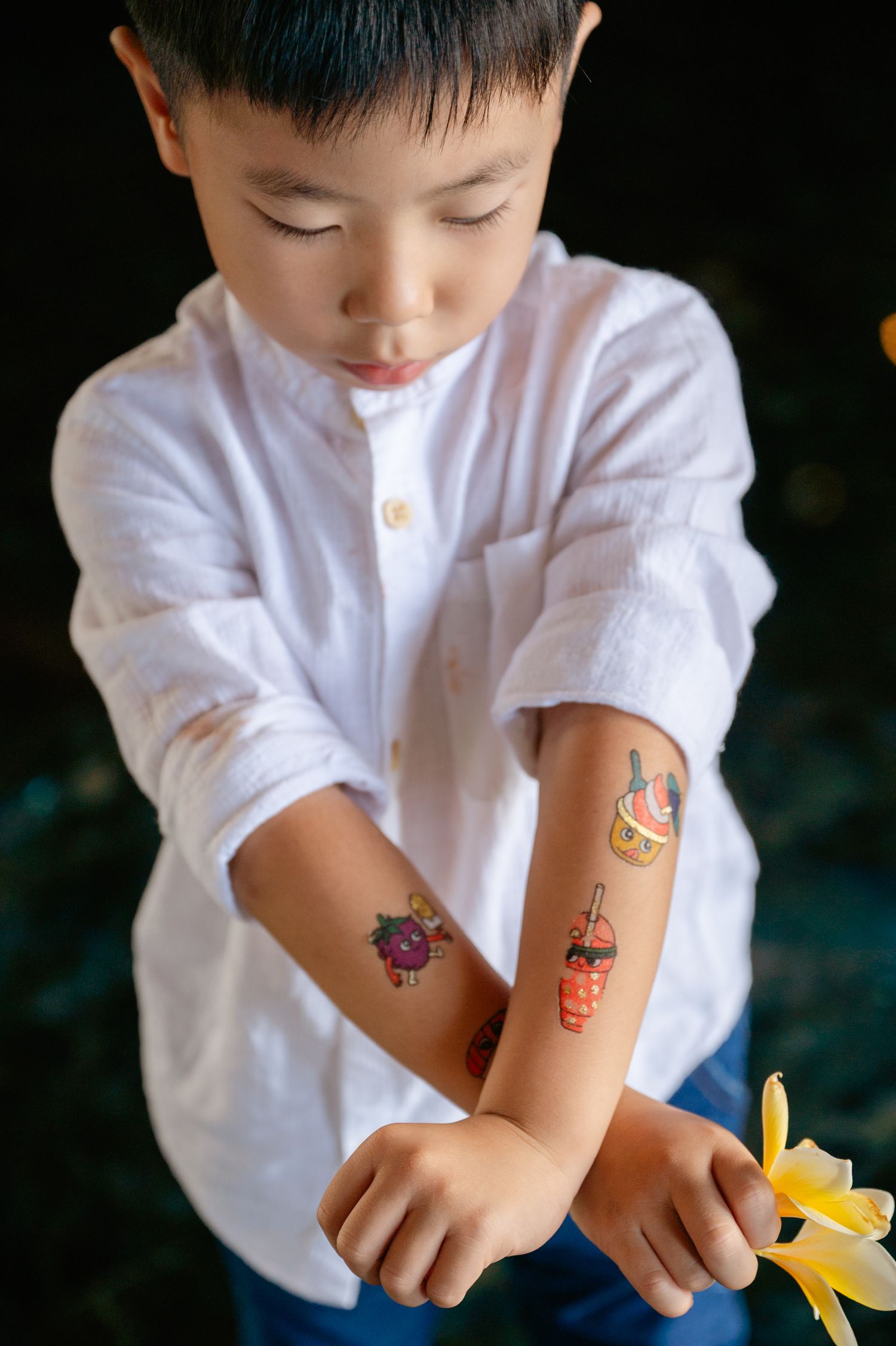 Young Asian boy in white shirt showing colorful temporary tattoos on his forearms outdoors.