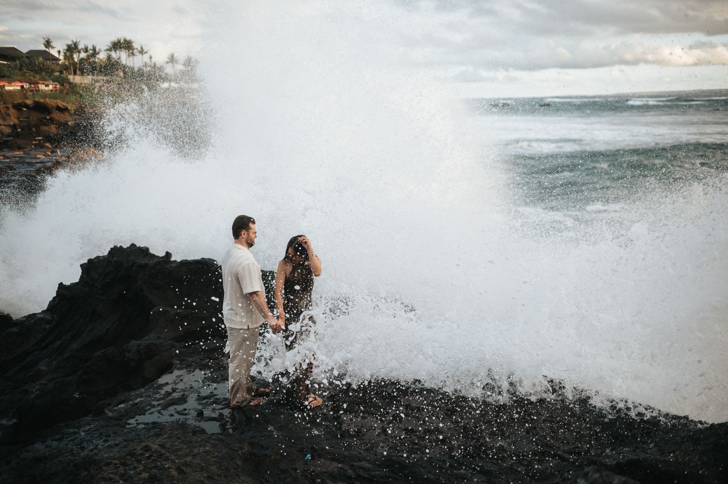Couple standing on rocky shore as massive ocean wave crashes behind them in Hawaii.