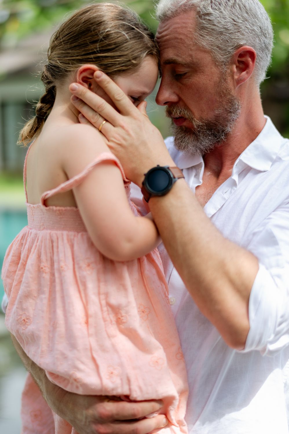 Elderly man with gray beard tenderly holds young girl in pink dress, touching her face in emotional embrace outdoors.