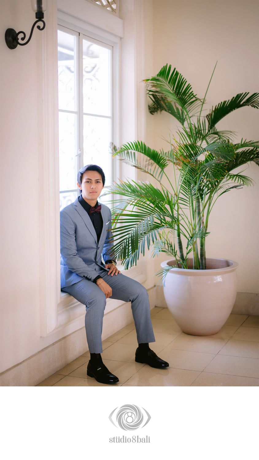 Young man in a light blue suit sitting by a bright window next to a large potted palm plant indoors.