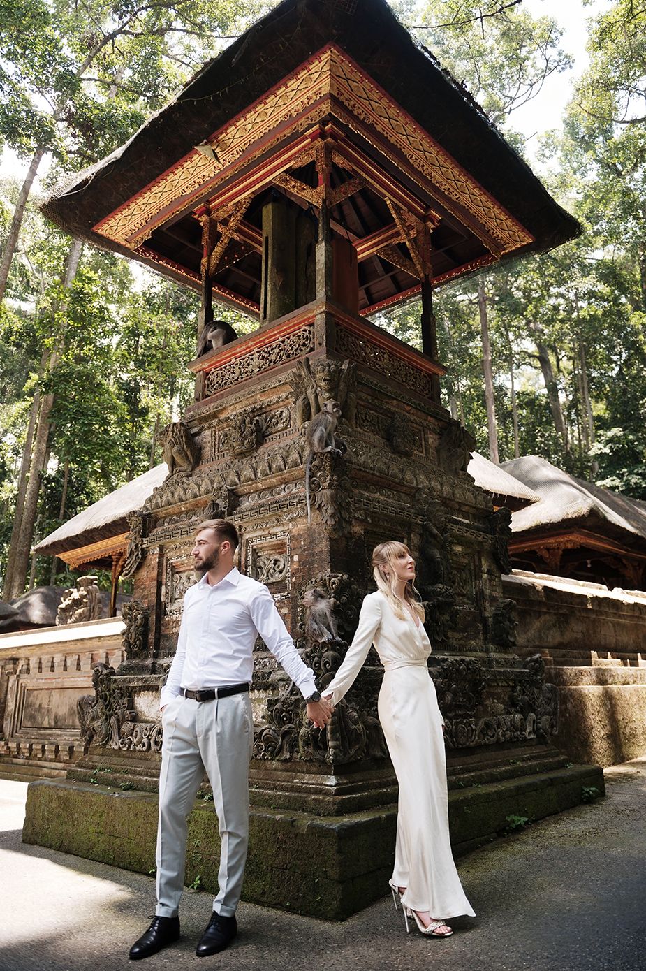 Couple holding hands in front of an ornate Balinese temple structure surrounded by lush green forest trees.