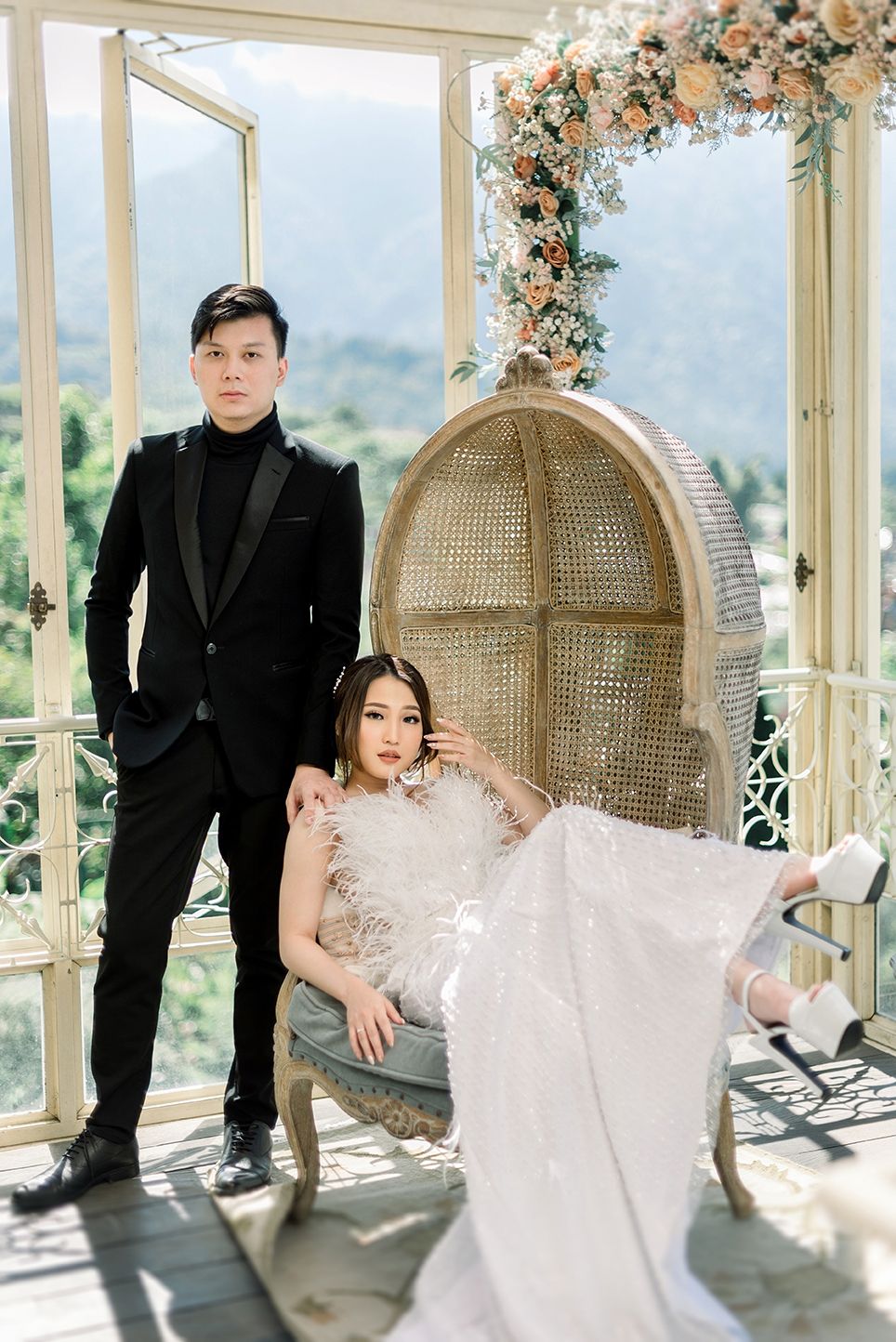 Couple in elegant wedding attire pose in a decorative rattan chair by open windows with floral arch and mountain views.