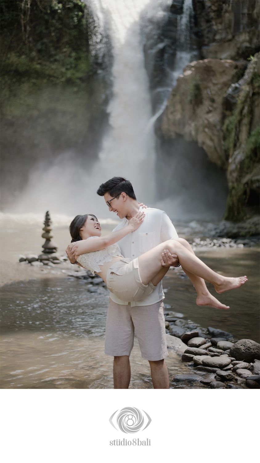 Couple posing romantically in front of a waterfall, man carrying woman in his arms on rocky shore in Bali.