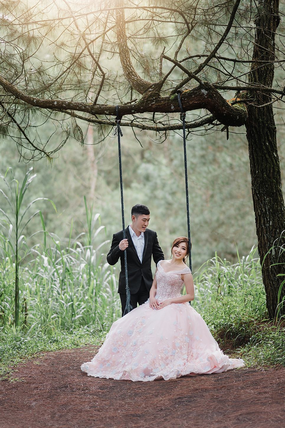 Couple in wedding attire pose by a tree swing in a lush forest, bride in pink gown, groom in black suit.