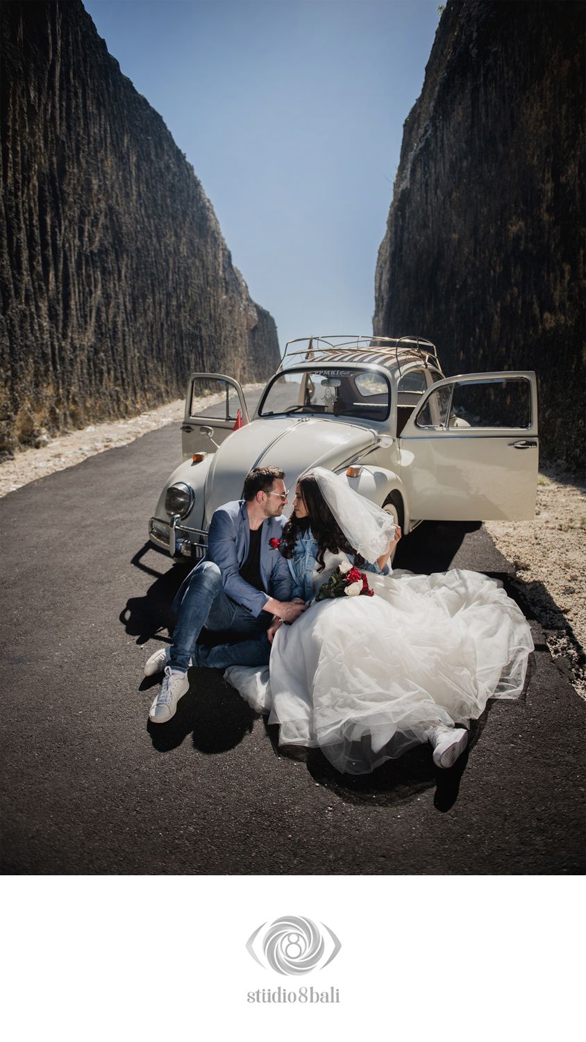 Bride and groom sitting together on a road beside a vintage white Volkswagen Beetle, framed by dramatic rocky cliffs.