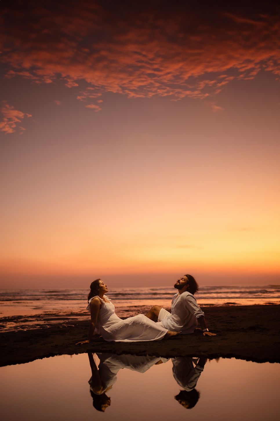 Couple in white sitting back-to-back on a beach at sunset, reflected in water under a dramatic orange sky.