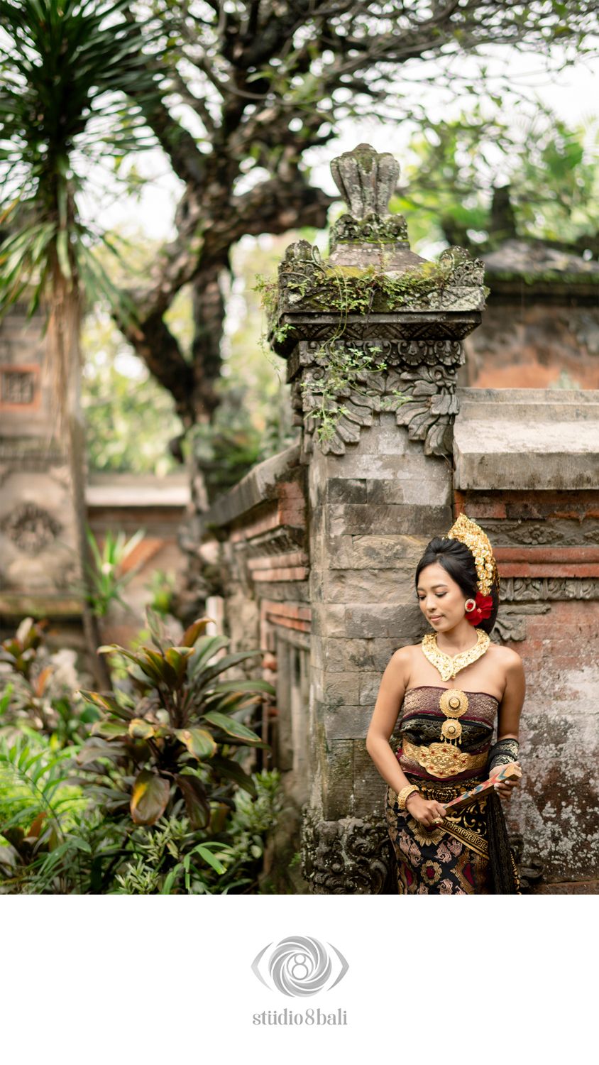 Balinese woman in traditional gold and batik kebaya dress poses by an ornate stone temple gate in Bali.