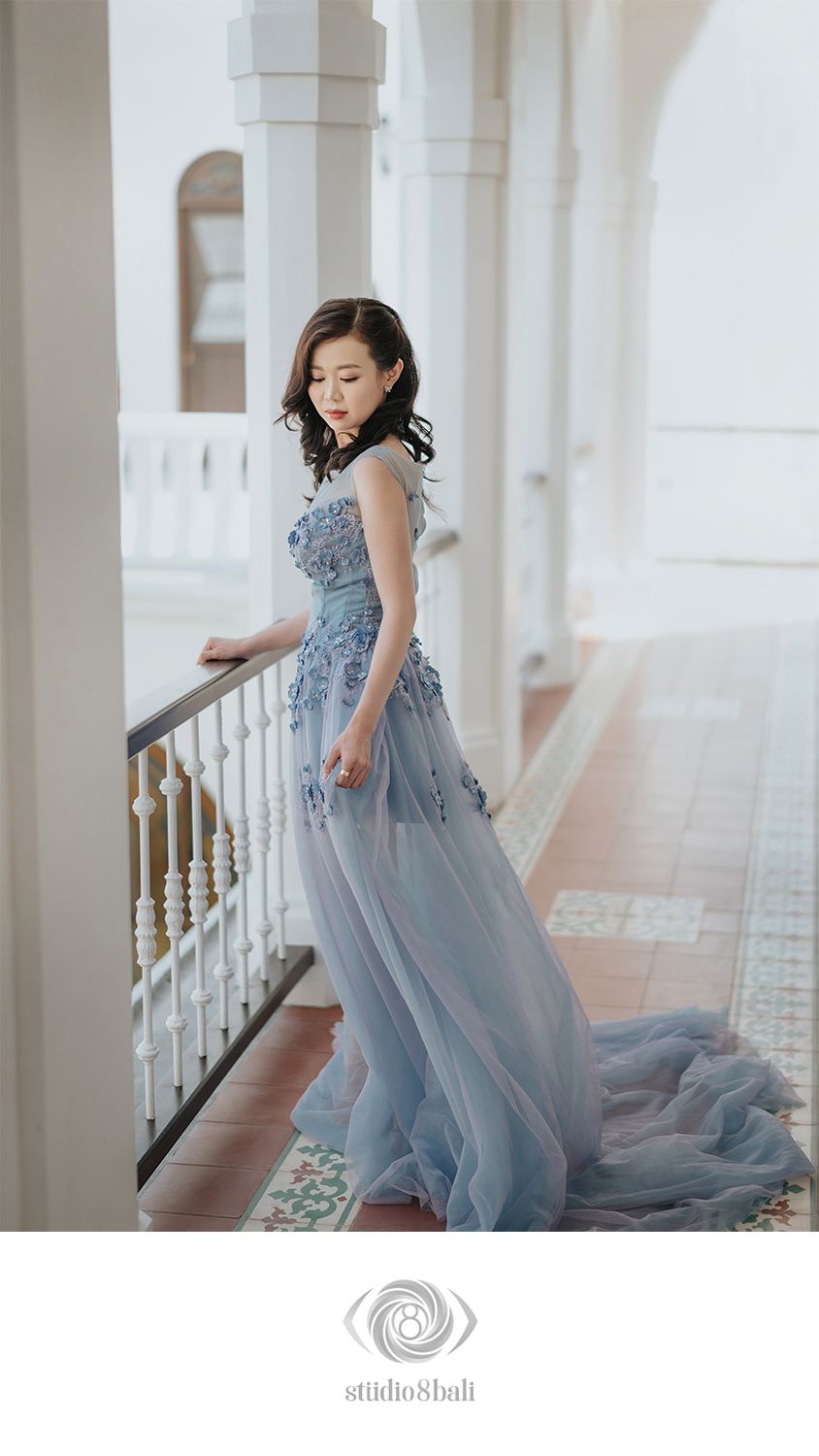 Woman in flowing blue-grey floral applique gown standing on ornate colonial balcony.