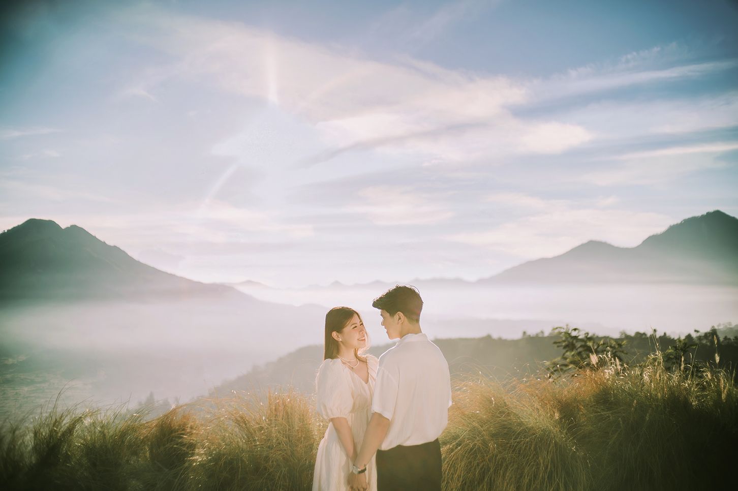 Couple sharing a romantic moment on a misty mountain summit at sunrise with scenic valley and mountain range backdrop.