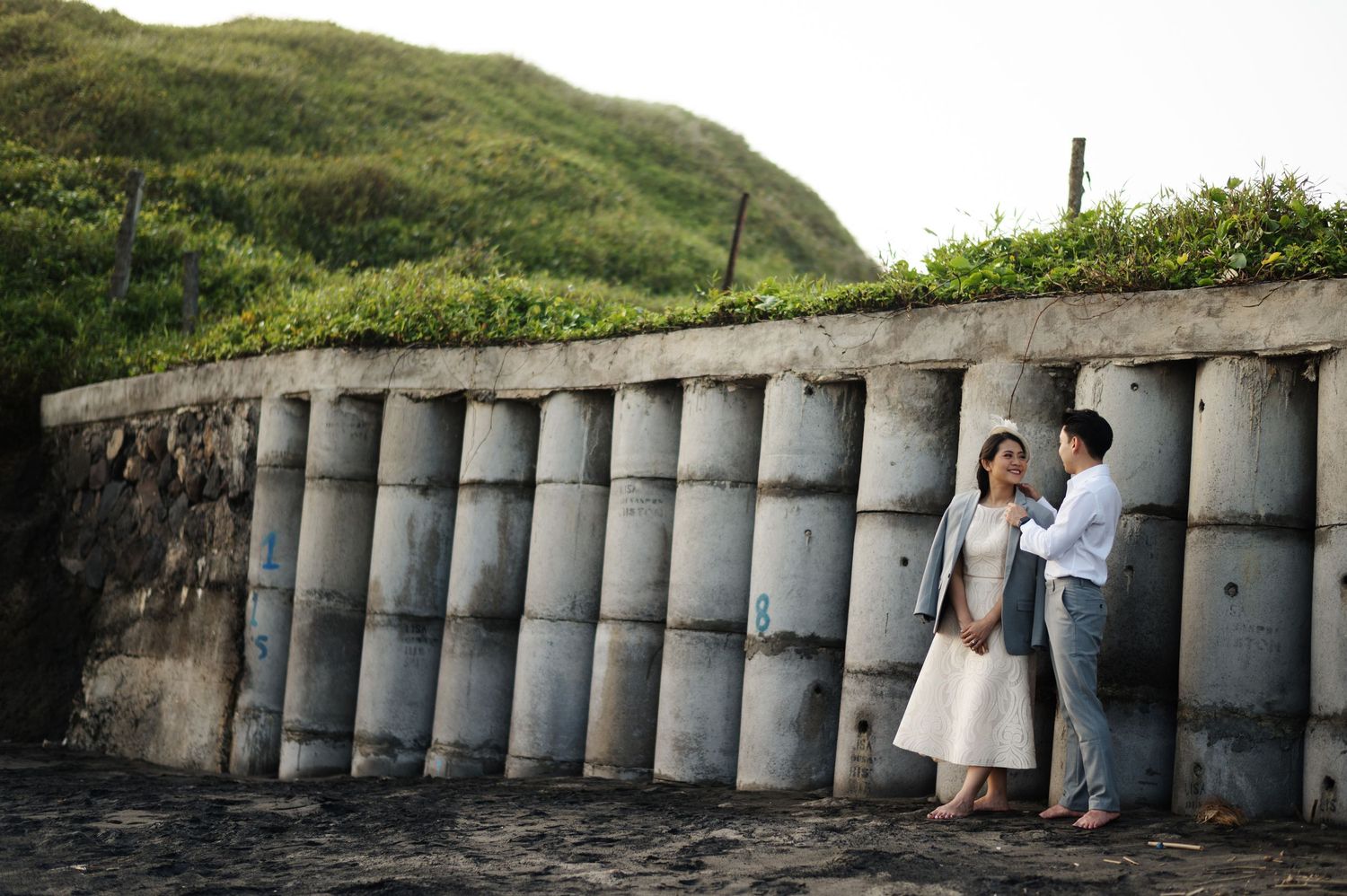 Couple sharing a romantic moment by a concrete sea wall with lush green hillside backdrop.