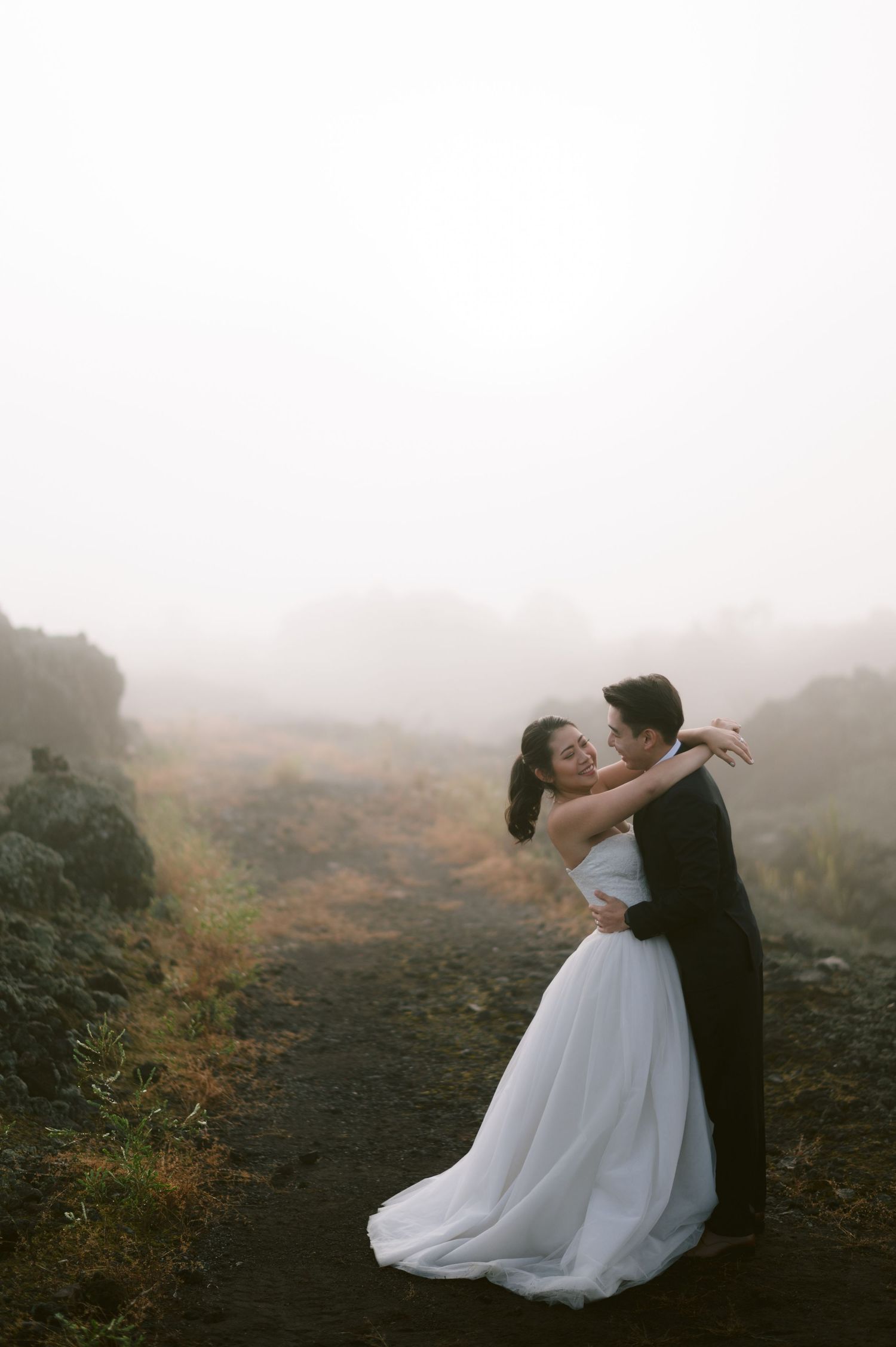 A bride and groom embrace romantically on a misty, foggy path, with the bride in a flowing white gown.