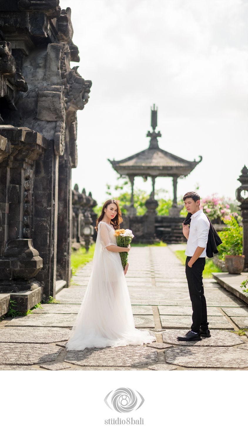 Couple in wedding attire posing at a Balinese temple pathway, bride in white gown holding bouquet, groom in black suit.