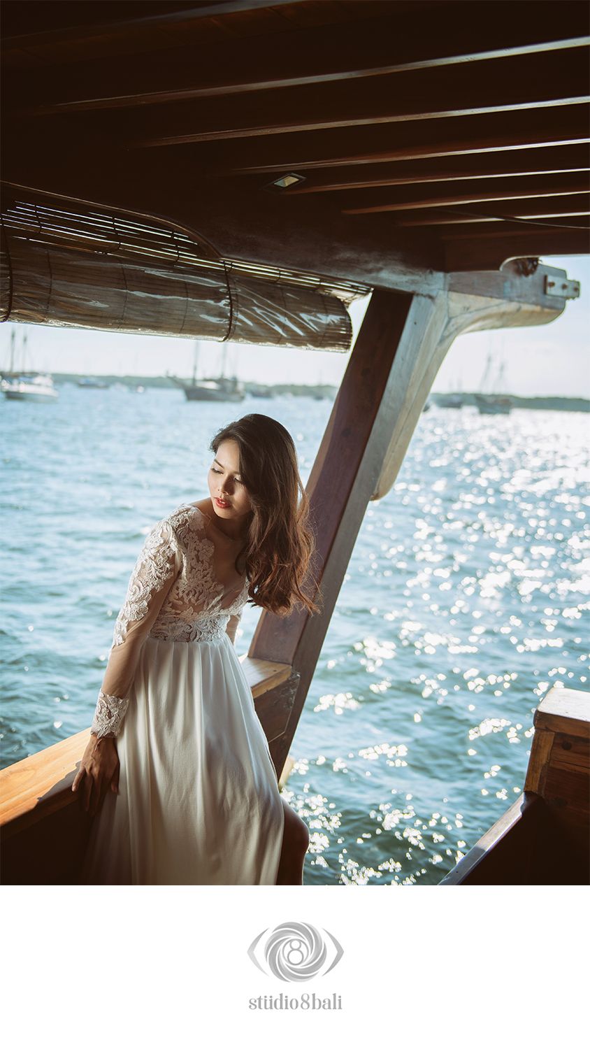 Bride in lace wedding gown standing on wooden boat with sparkling ocean behind her.