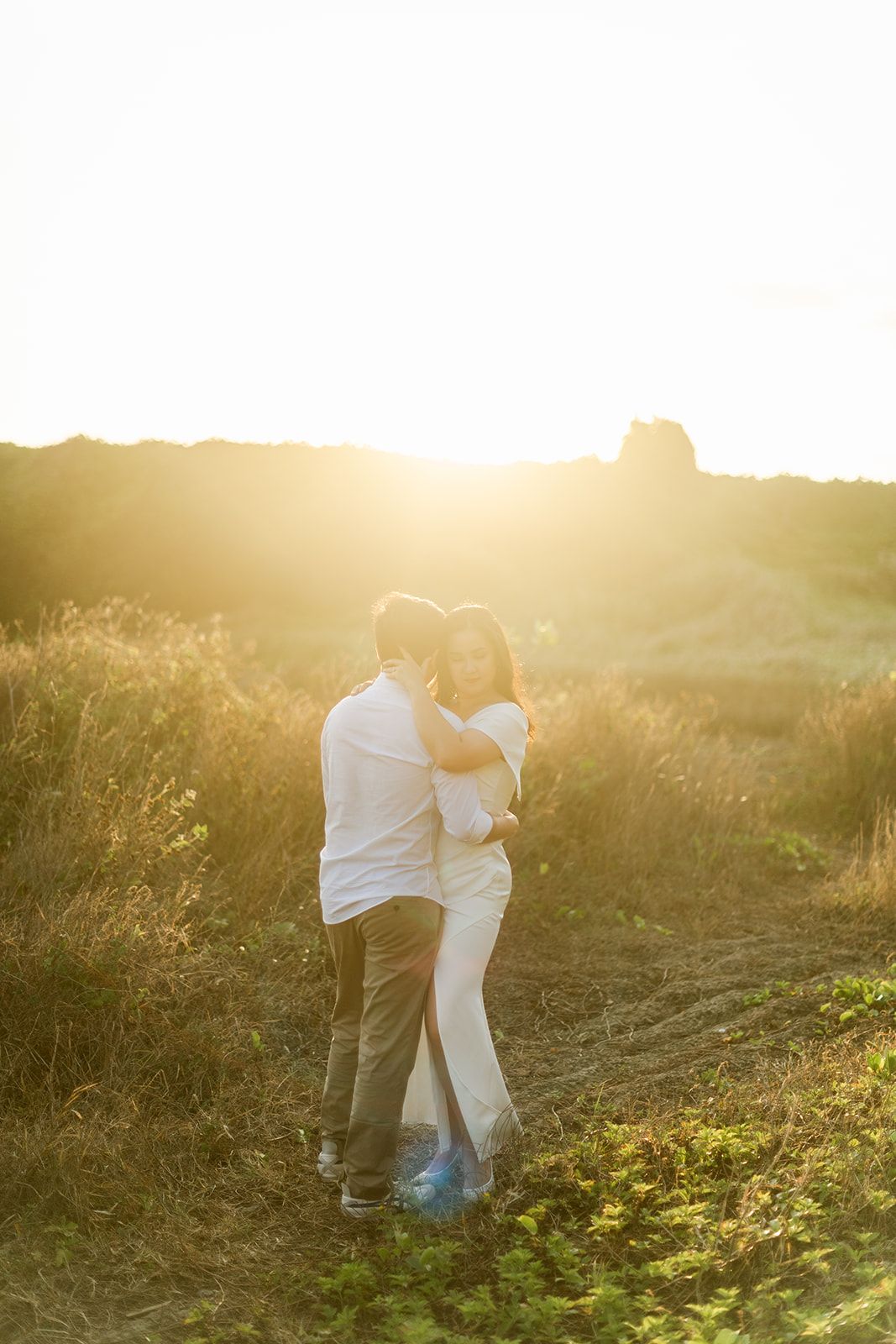 Couple embracing in golden sunset light in a grassy field, romantic outdoor photography with warm backlight.