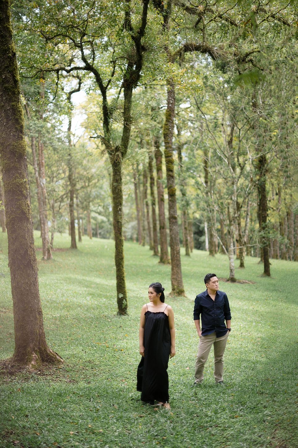 Couple in black attire standing apart in a lush green forest with tall moss-covered trees.