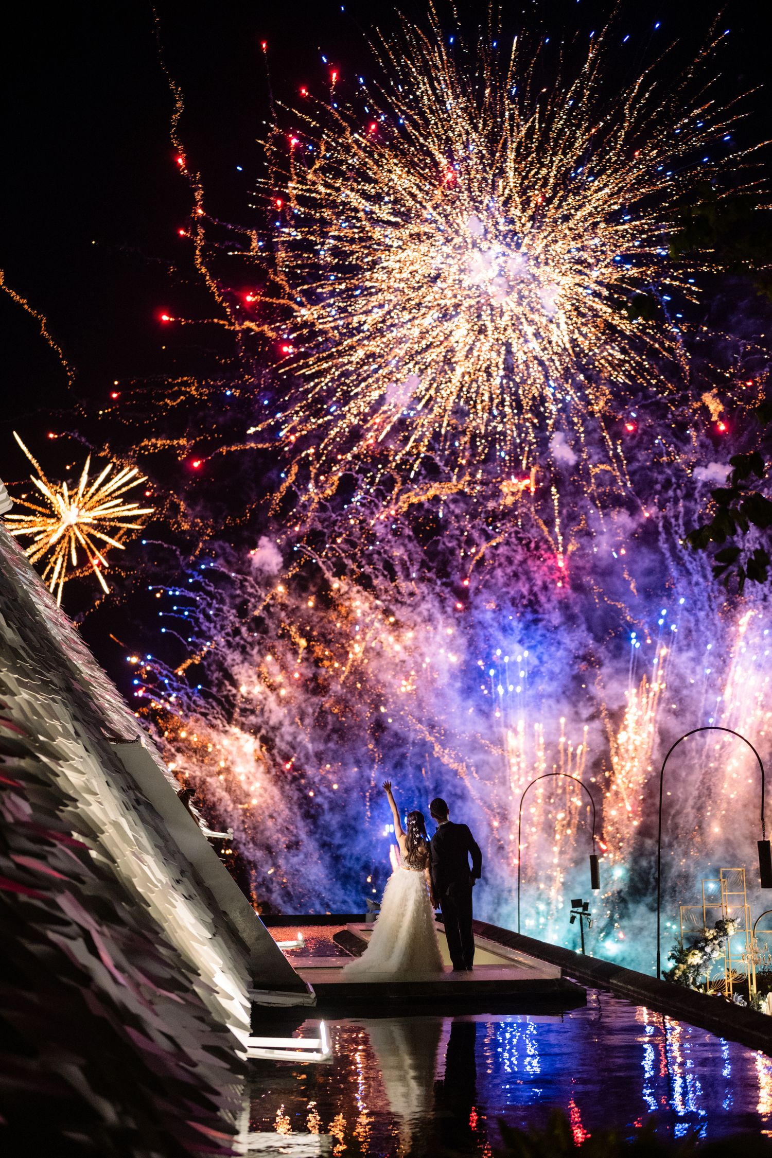 Bride and groom stand beside a reflecting pool at night as colorful fireworks burst across the dark sky above them.
