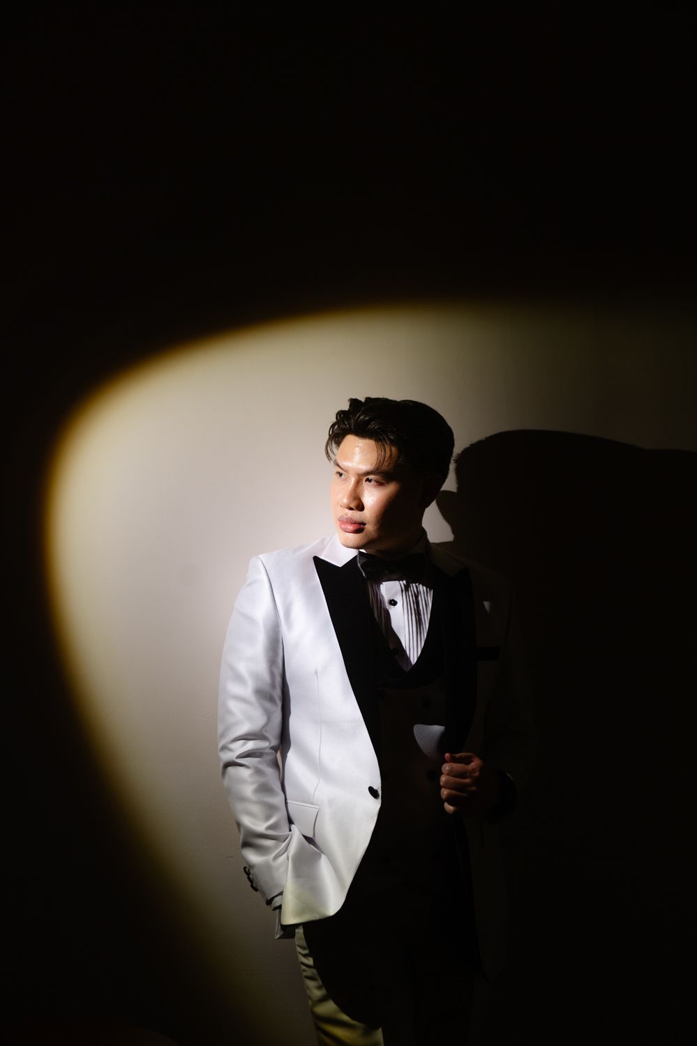 Young Asian man in white tuxedo jacket and black bow tie posed dramatically under spotlight against dark background.
