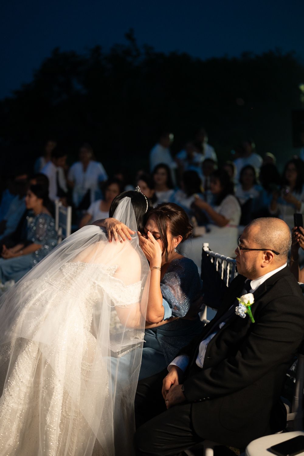 Bride in white gown and veil embracing emotional guest at outdoor evening wedding ceremony.