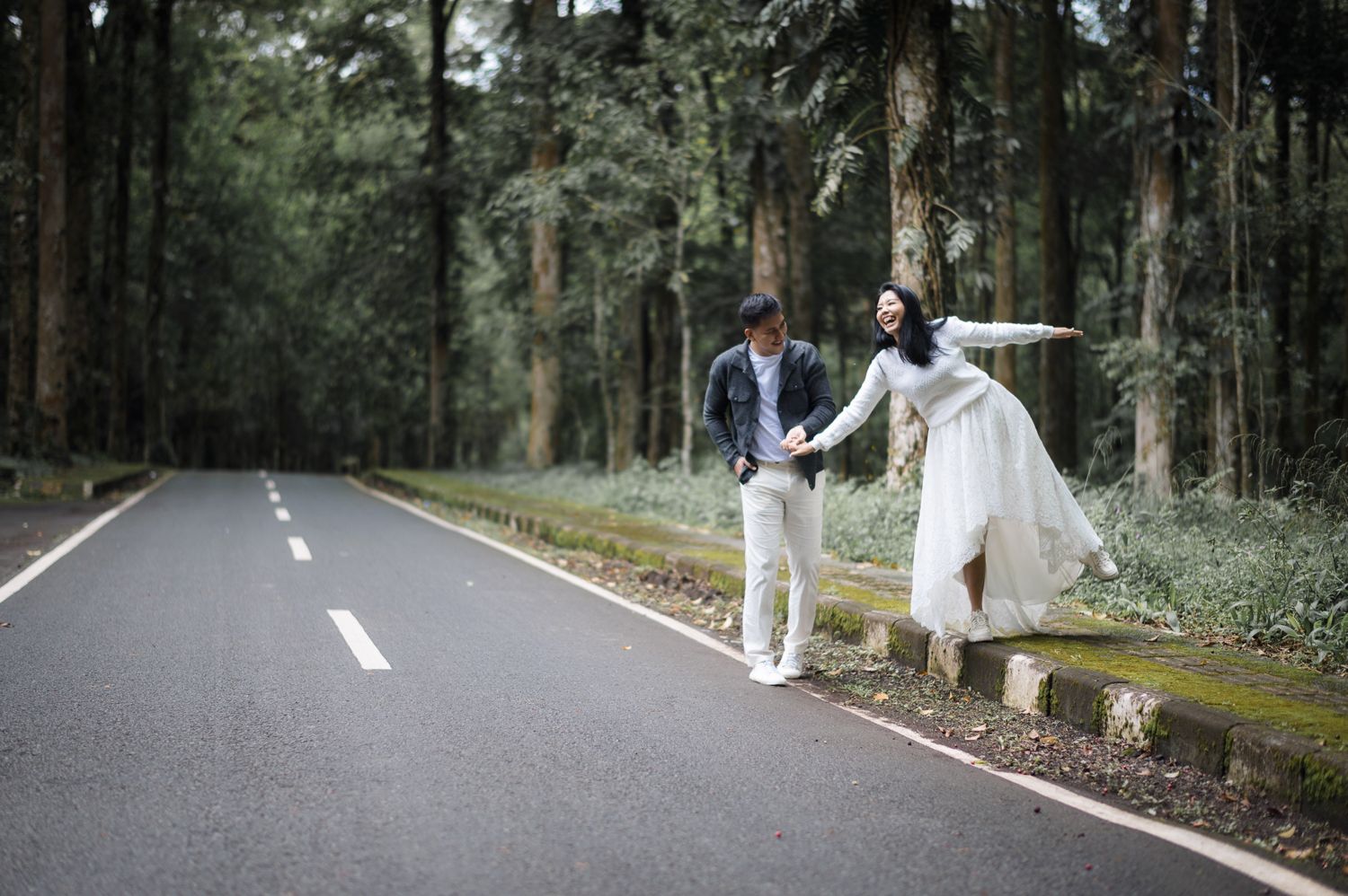 Couple in wedding attire holding hands on a tree-lined road, bride in white gown with arms outstretched.