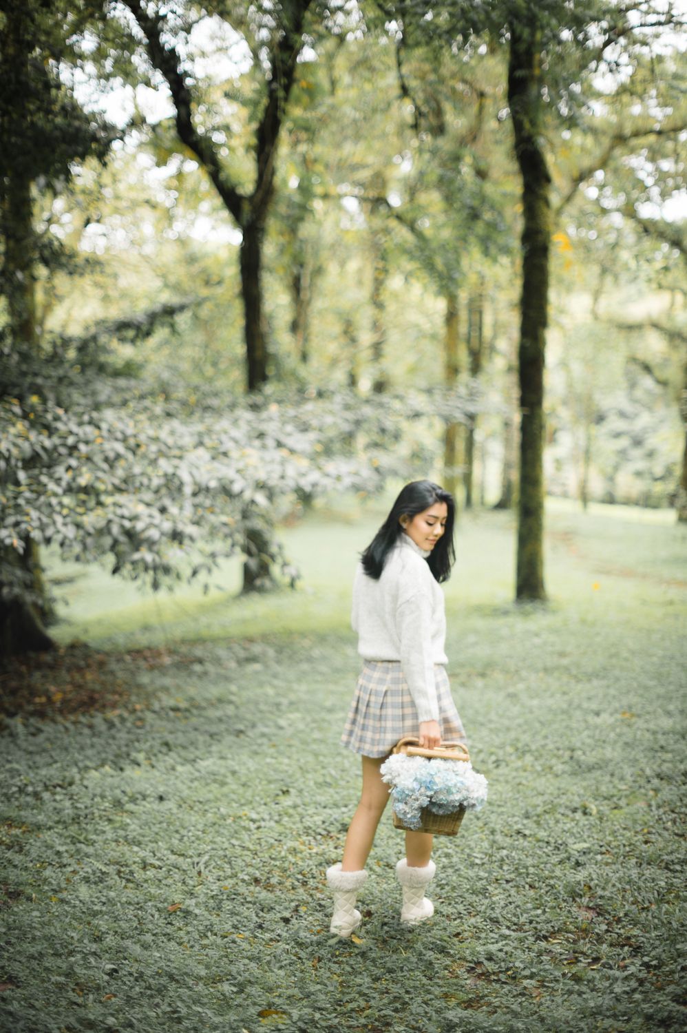 Young woman in plaid skirt and white sweater walks through lush green forest carrying a bouquet of blue flowers.
