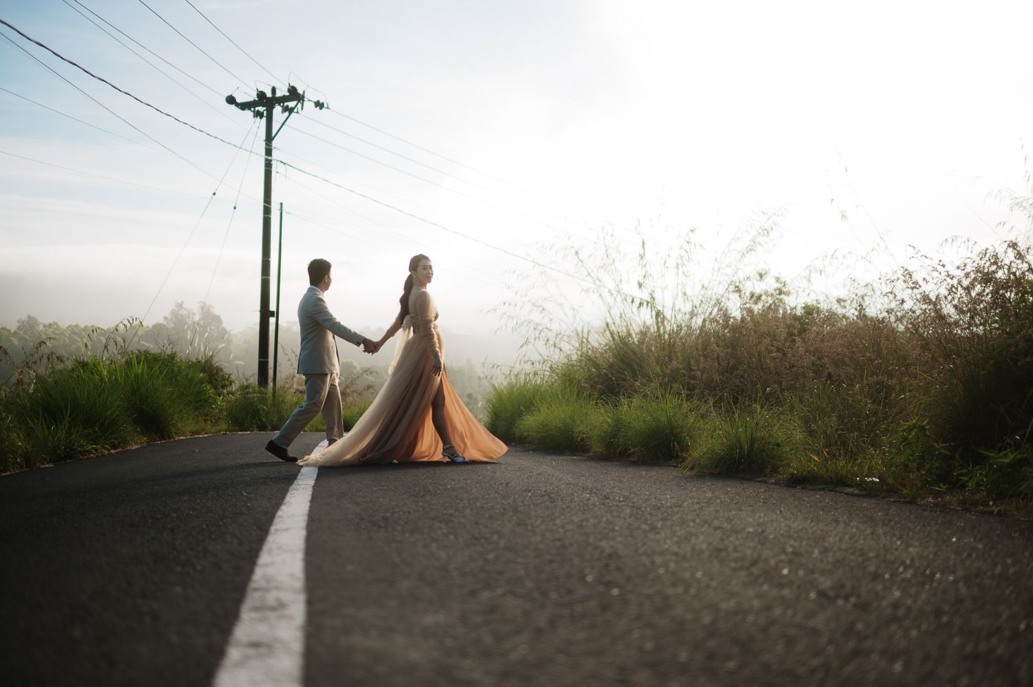 Bride and groom holding hands walking on a misty road at sunrise.