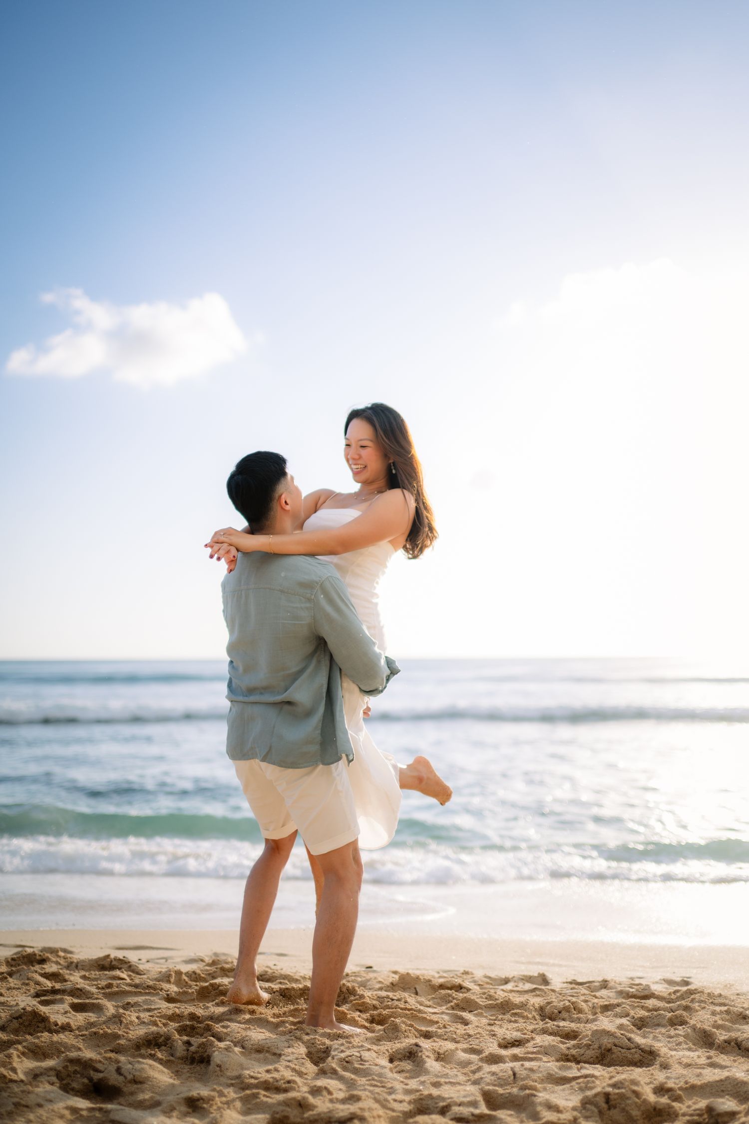 A couple embraces on a sandy beach, the man lifting the smiling woman as ocean waves and blue sky fill the background.