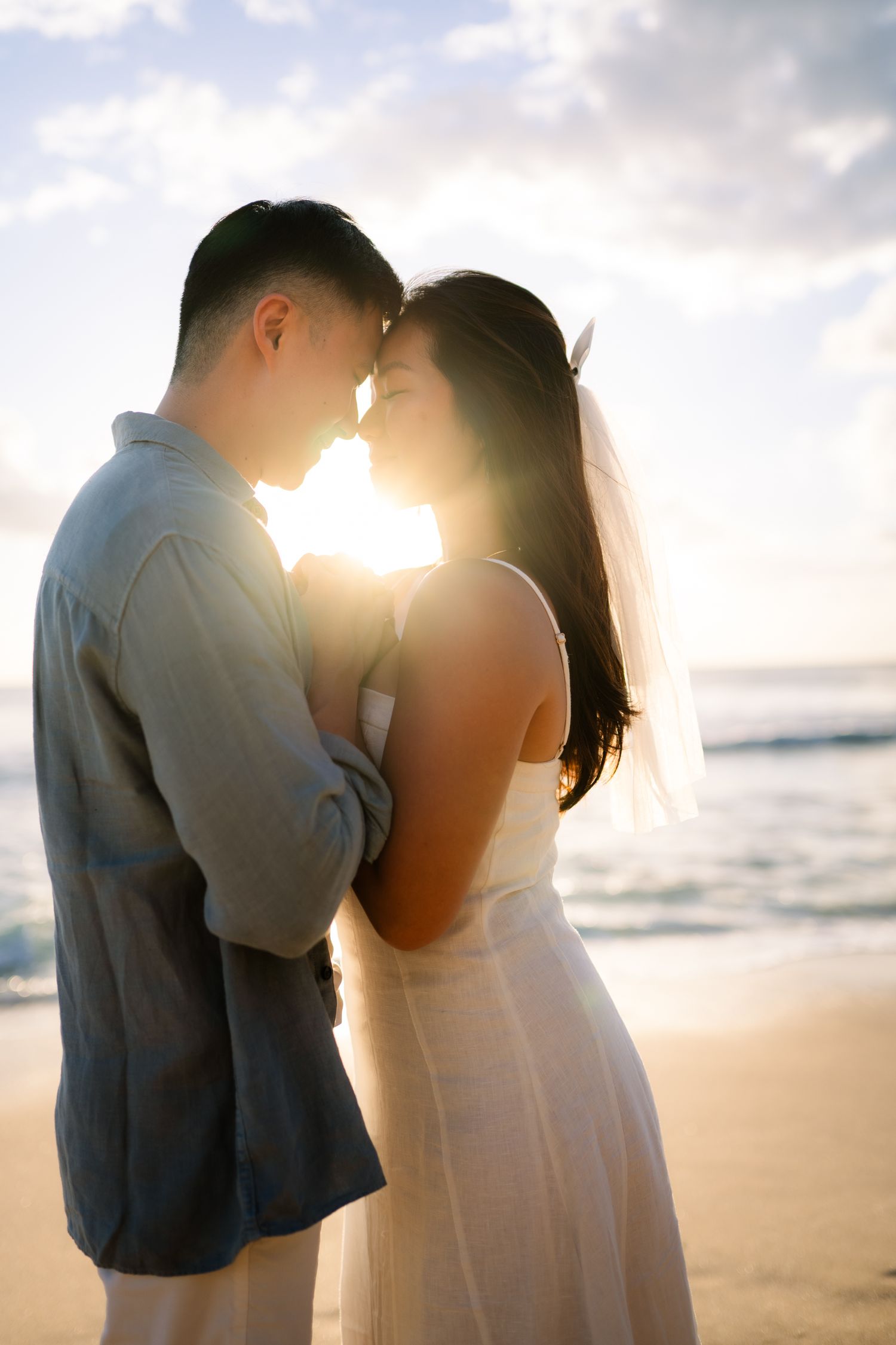 Bride and groom share a romantic sunset beach moment, foreheads touching, with ocean waves in the background.