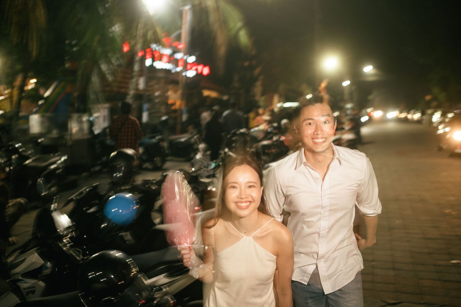 Young couple smiling and walking at night near parked motorcycles, woman holding pink cotton candy.
