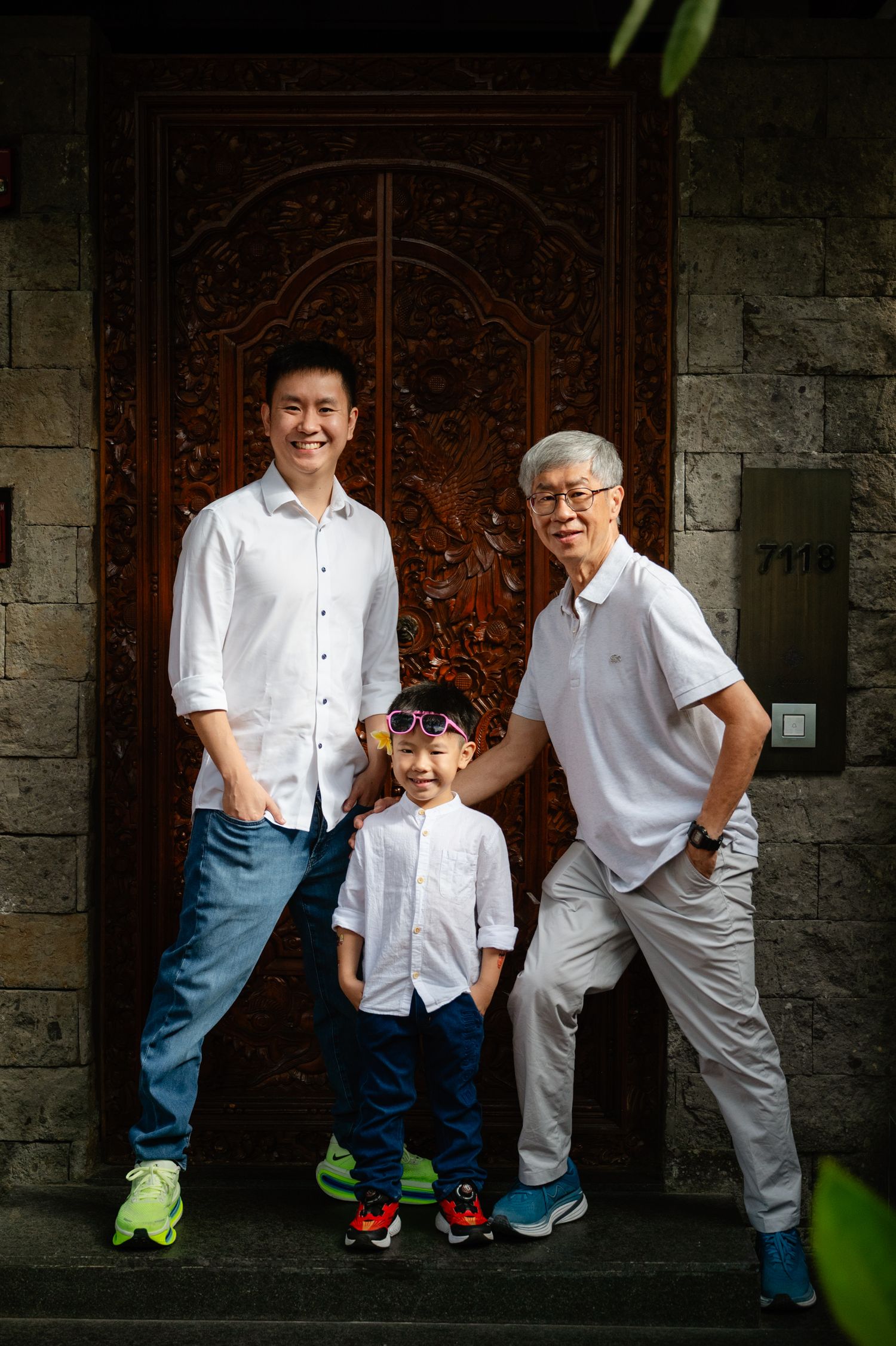 Three generations of men pose smiling before ornate wooden doors, with a young boy wearing colorful sunglasses.