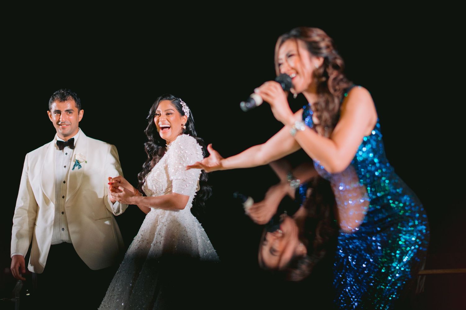Bride and groom dancing joyfully at wedding reception while female singer in blue sequin gown performs on stage.